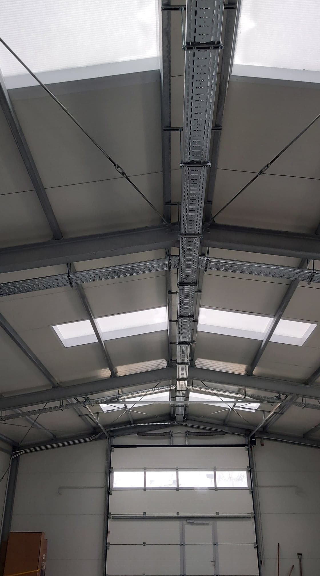 Wnętrze hali przemysłowej z widoczną konstrukcją stalową dachu, oświetleniem naturalnym przez okna dachowe oraz korytkami kablowymi zamontowanymi pod sufitem.