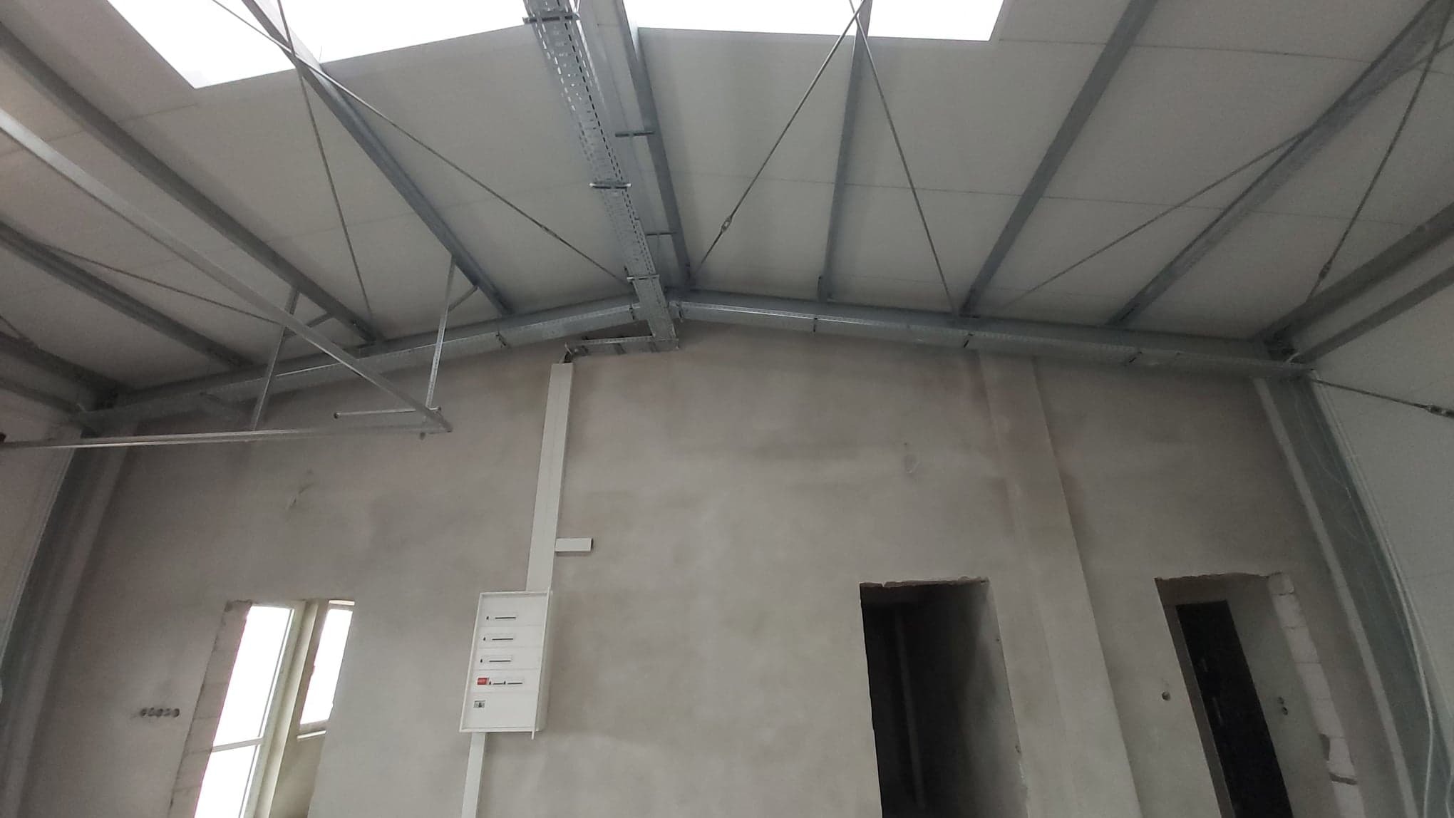 Wnętrze hali w stanie surowym, widoczne metalowe elementy konstrukcji dachu, koryta kablowe pod sufitem, biała skrzynka elektryczna na ścianie, otwory drzwiowe w betonie.