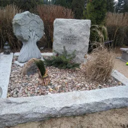 Granitowy nagrobek z inskrypcją i rzeźbą drzewa, otoczony kamieniami, z lampionami i roślinami na cmentarzu.