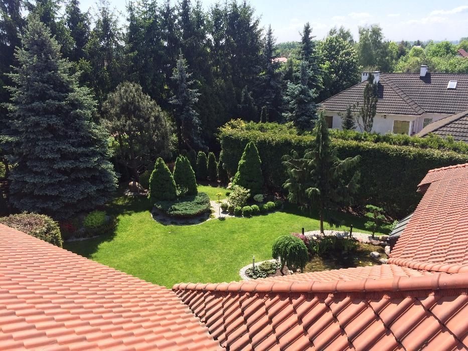 Widok z góry na zadbany ogród z równo przystrzyżonym trawnikiem, ozdobnymi krzewami i oczkiem wodnym, otoczony wysokimi drzewami i żywopłotem, z widocznym fragmentem dachu domu.