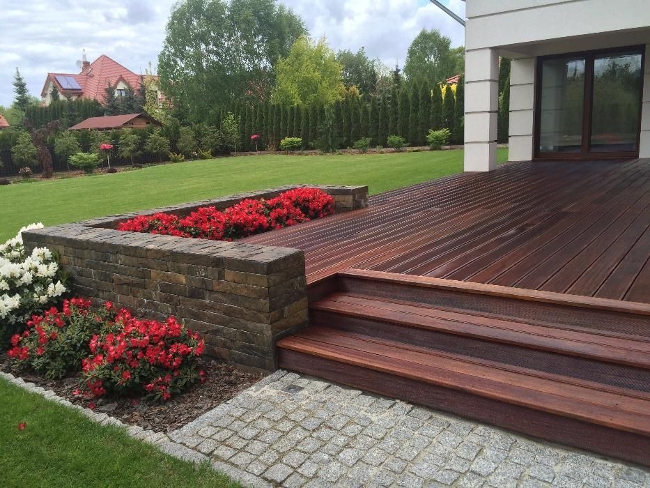 Ogród z tarasem z ciemnego drewna, schodami i rabatami kwiatowymi z czerwonymi i białymi kwiatami, obłożonymi kamieniami, na tle zadbanego trawnika i domu.