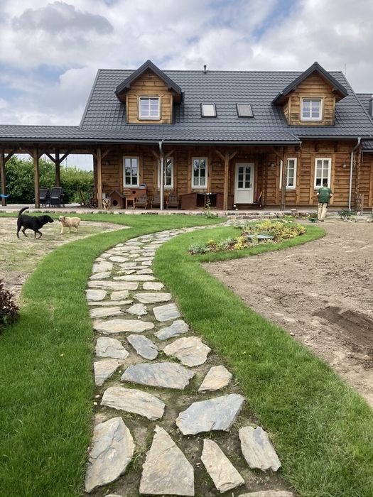 Kręta ścieżka z nieregularnych kamieni prowadząca do drewnianego domu z ciemnym dachem, trawnik świeżo założony, dwa psy biegające po podwórku, osoba w ogrodzie w tle.