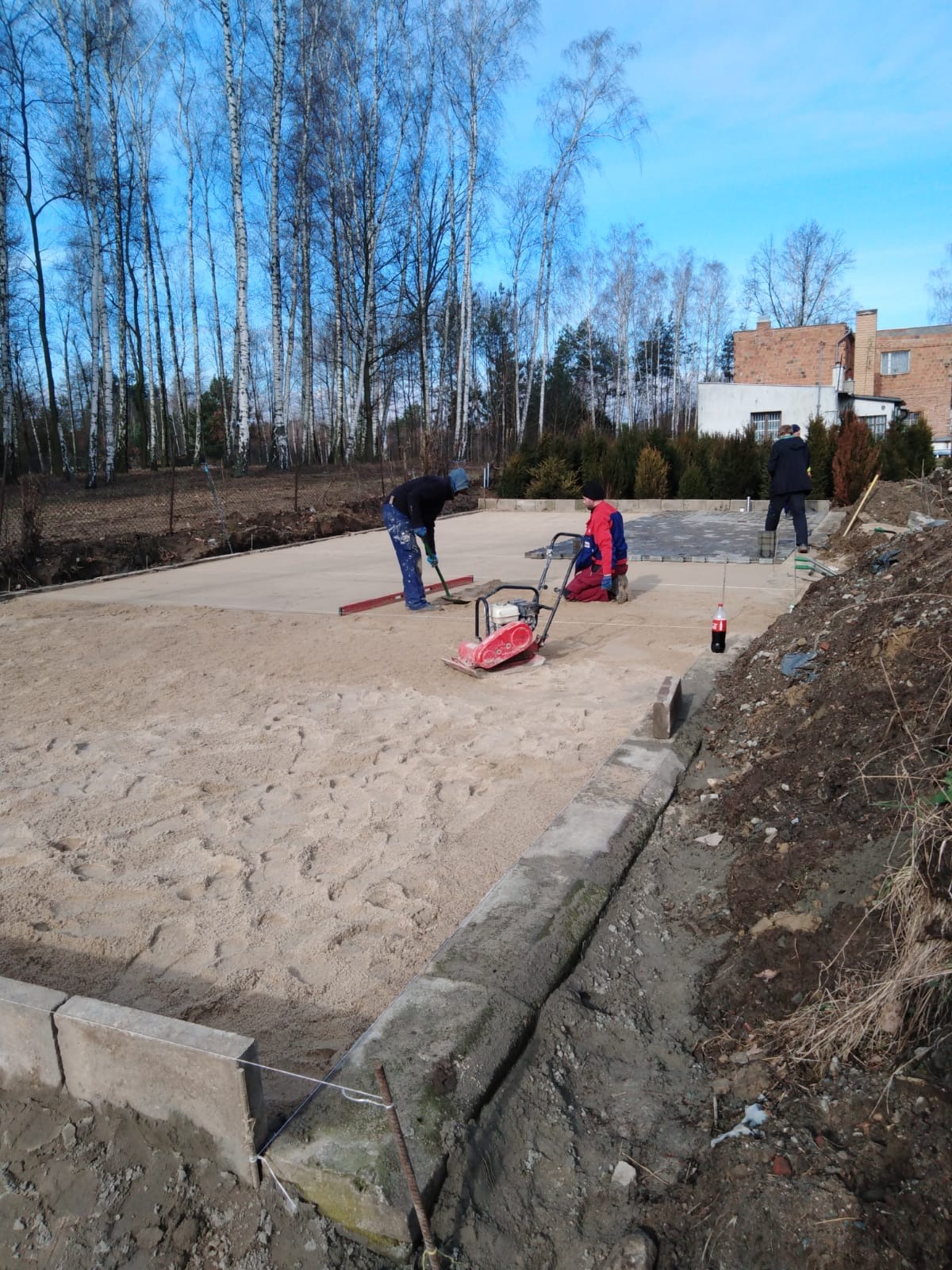 Prace przygotowawcze pod nawierzchnię z kostki: wyrównywanie piasku, ubijanie zagęszczarką, widoczne obrzeża i częściowo ułożona kostka w tle, robotnicy w trakcie pracy.