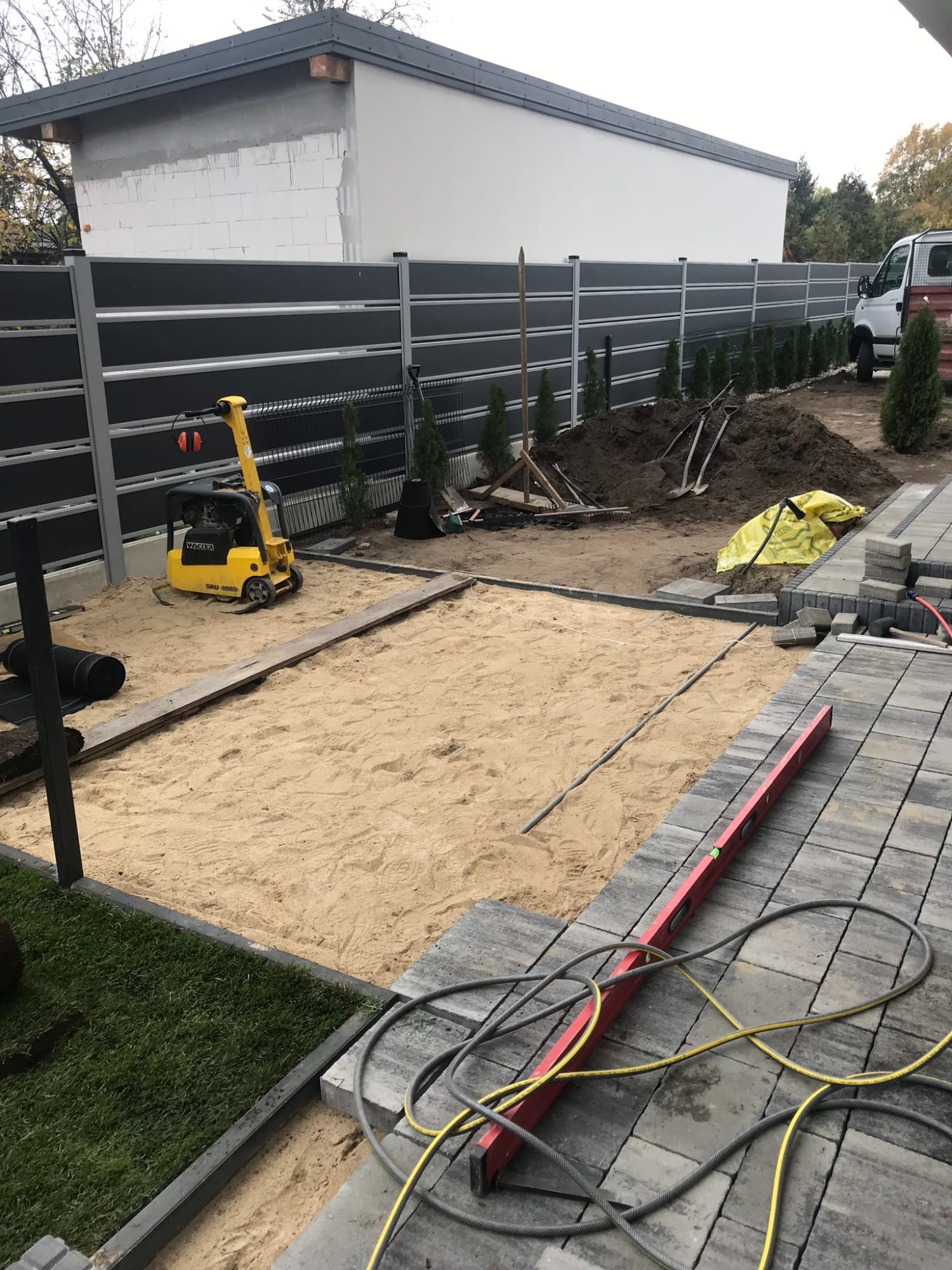 Przygotowanie podłoża pod nawierzchnię z kostki brukowej: ubijarka wibrująca na piasku, częściowo ułożona kostka, poziomica, w tle ogrodzenie i małe drzewka.