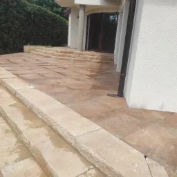 Jasne, kwadratowe płyty chodnikowe ułożone w schody i chodnik przed budynkiem z ciemnymi drzwiami wejściowymi i czarną rynną spustową, widoczne ślady wilgoci.