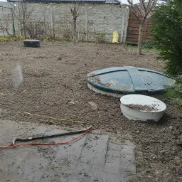 Montaż przydomowej oczyszczalni ścieków: widoczne dwa włazy do zbiorników w wykopanym gruncie, z narzędziem (łom) i kablem na pierwszym planie, oraz betonowe ogrodzenie panelowe i dom z panelami...