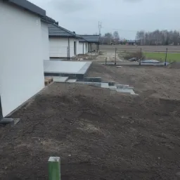 Przygotowanie terenu pod budowę w Wrześni: wyrównana ziemia, widoczne fundamenty domów i ogrodzenie panelowe w tle.