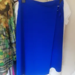 Kobaltowa spódnica kopertowa z wiązaniem na boku, wisząca na wieszaku obok innych ubrań w garderobie.