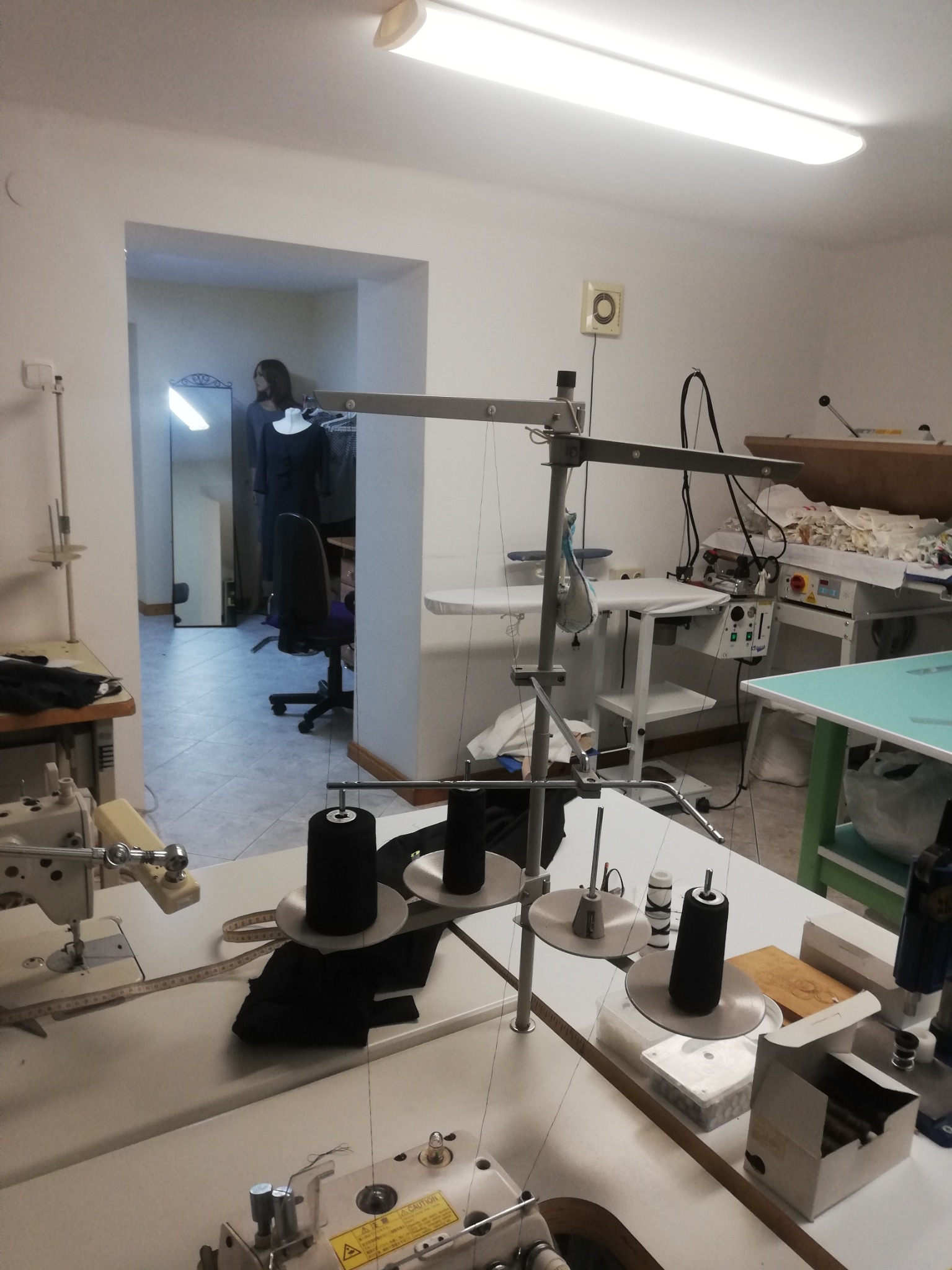 Wnętrze pracowni krawieckiej z kilkoma maszynami do szycia, stołami, żelazkiem przemysłowym i widoczną w głębi sylwetką osoby przy lustrze.