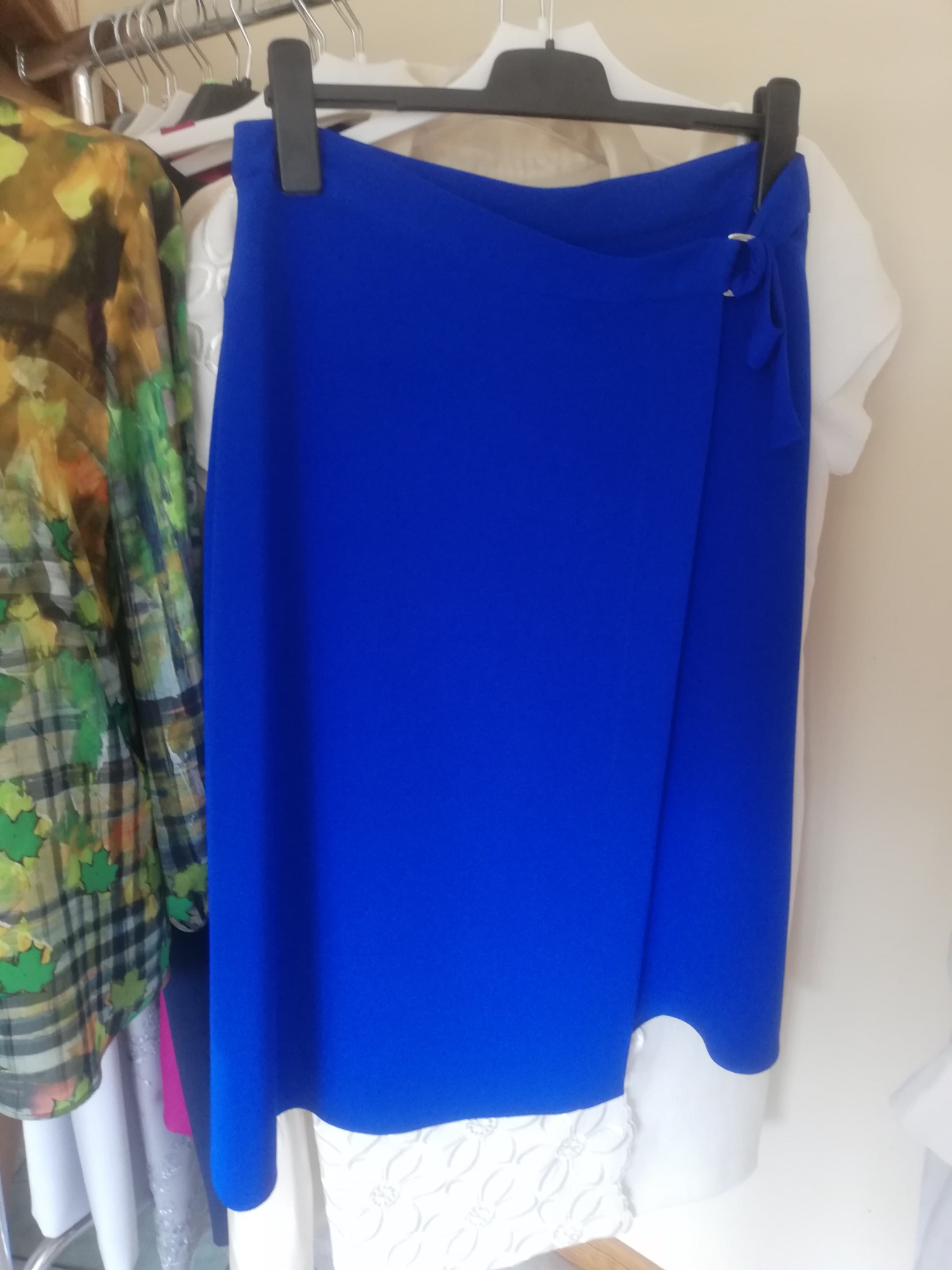 Kobaltowa spódnica kopertowa z wiązaniem na boku, wisząca na wieszaku obok innych ubrań w garderobie.