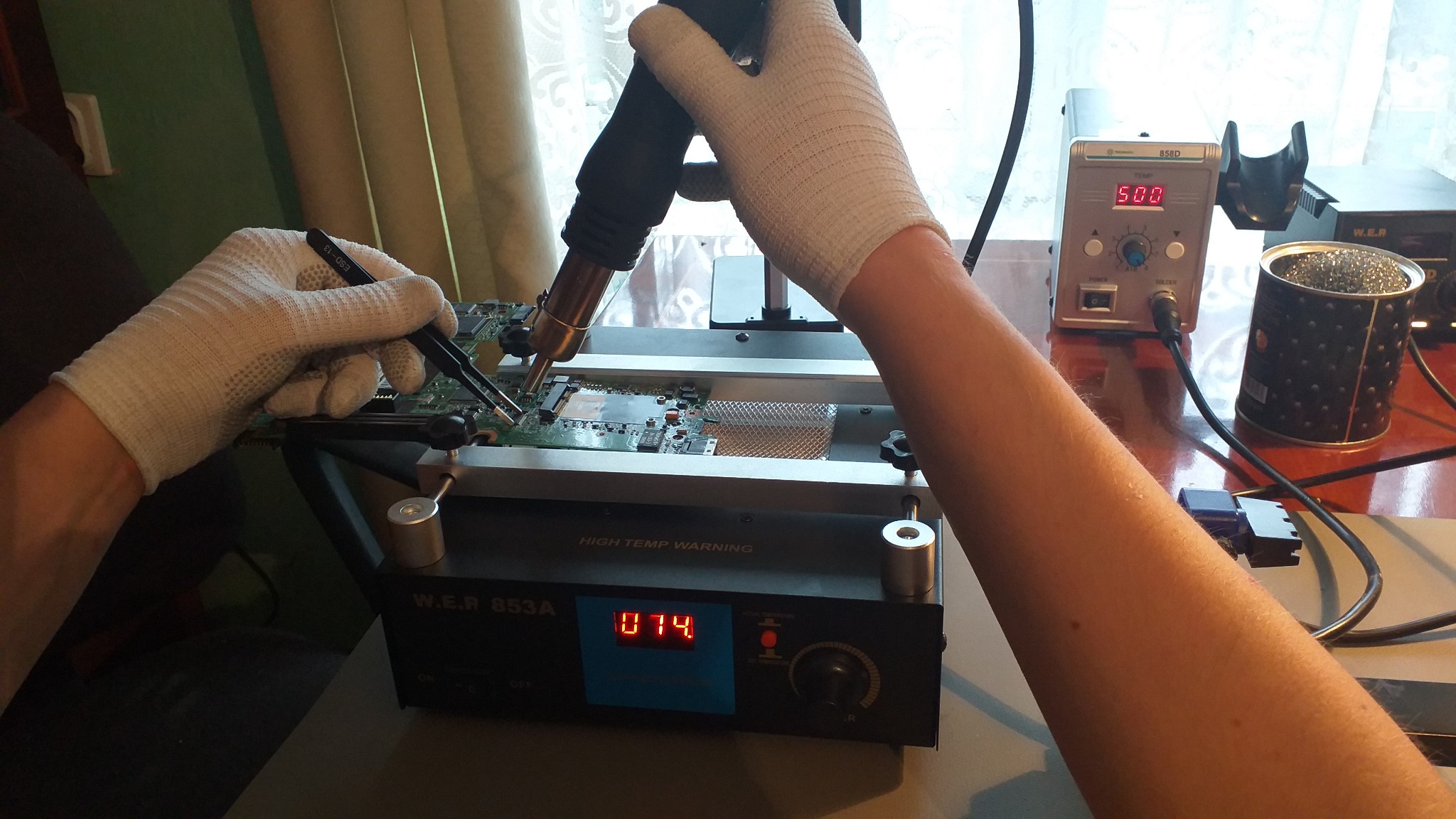 Serwisant w Legnicy w rękawicach ochronnych lutuje płytę główną komputera za pomocą pęsety i hot air rework station. Na stole widoczna stacja lutownicza z wyświetlaczem temperatury.