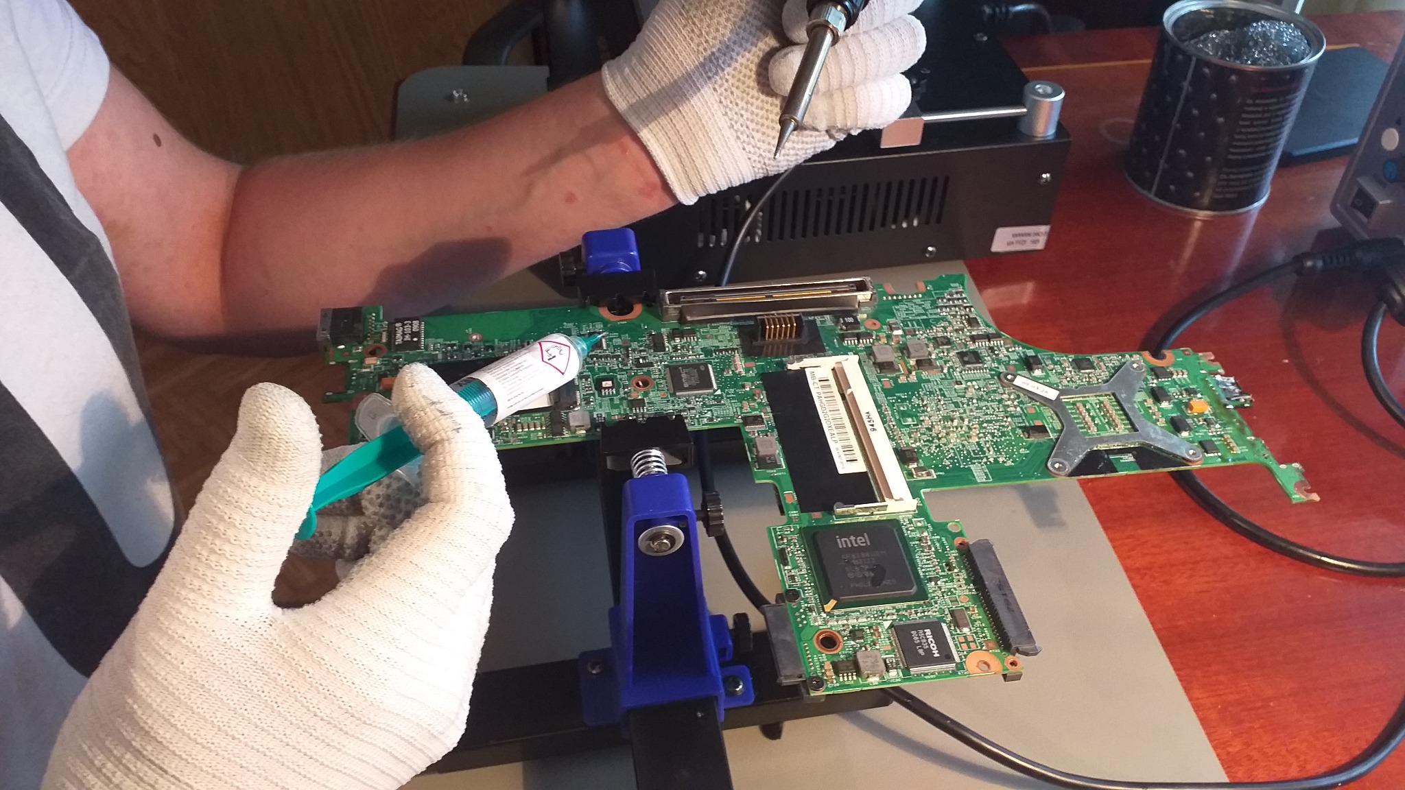 Serwisant w rękawicach antystatycznych aplikuje pastę termoprzewodzącą na płytę główną laptopa zamocowaną w uchwycie, obok lutownica i akcesoria.