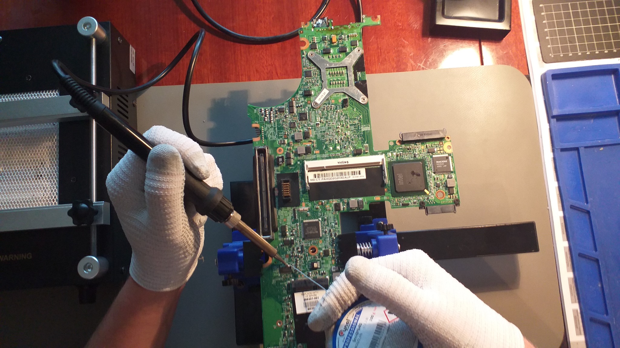 Lutowanie płyty głównej laptopa w rękawicach ochronnych, widoczne narzędzia: lutownica, topnik, stacja lutownicza, płyta zamocowana w uchwytach.