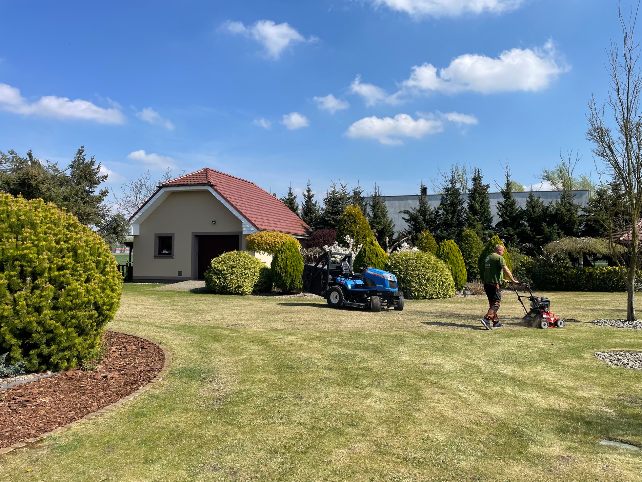 Mężczyzna kosi trawę kosiarką spalinową na tle zadbanego ogrodu z domem, krzewami i niebieskim traktorkiem do koszenia