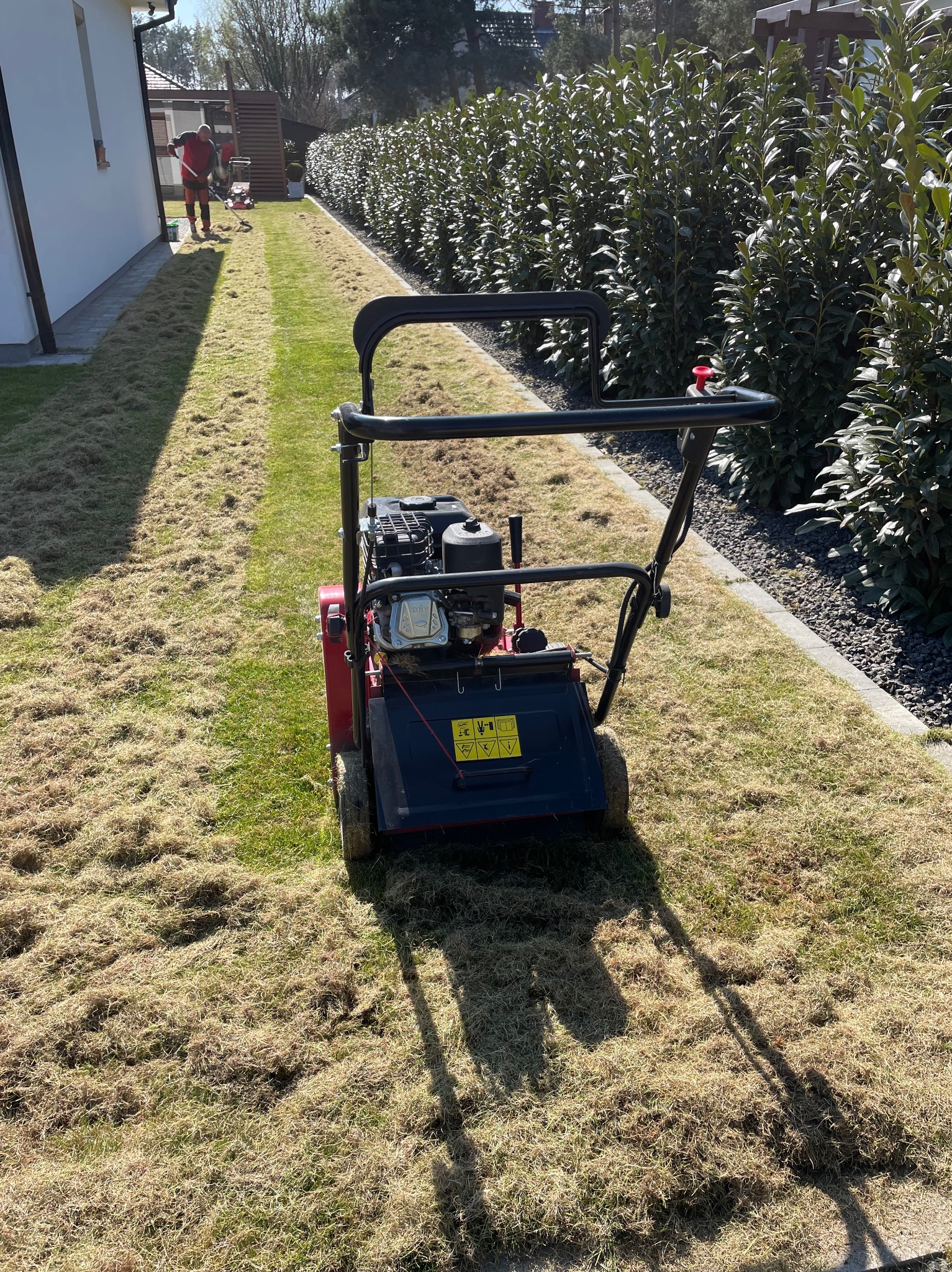 Wertykulator stojący na trawniku po wertykulacji, z widocznymi resztkami organicznymi. W tle osoba grabiąca trawnik wzdłuż białego budynku i żywopłotu.