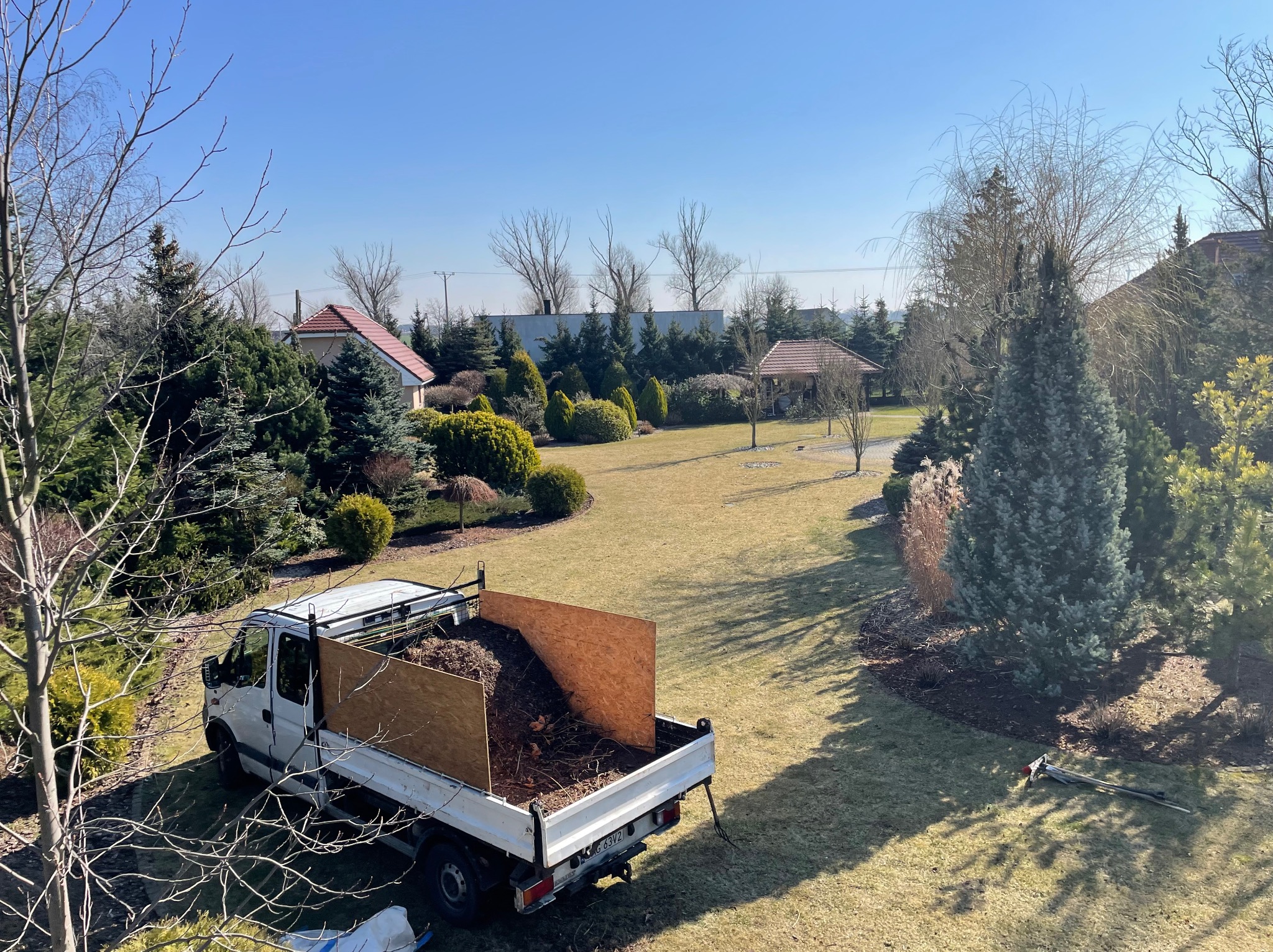 Widok z góry na zadbany ogród z podjazdem i białym samochodem dostawczym z przyczepką wypełnioną gałęziami i liśćmi, stojącym na trawniku.