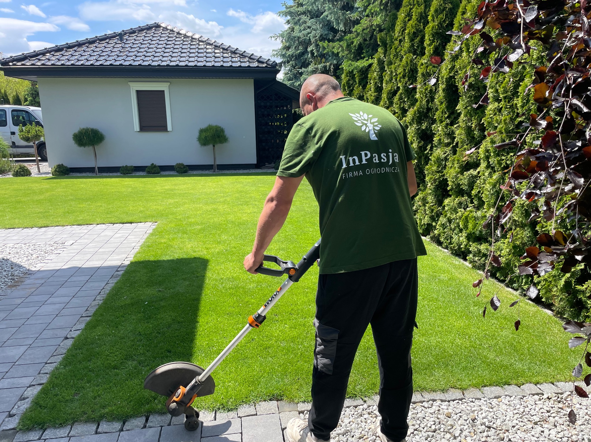 Mężczyzna w koszulce firmy ogrodniczej przycina trawę pod krzewami, używając podkaszarki elektrycznej na tle zadbanego trawnika i nowoczesnego domu.