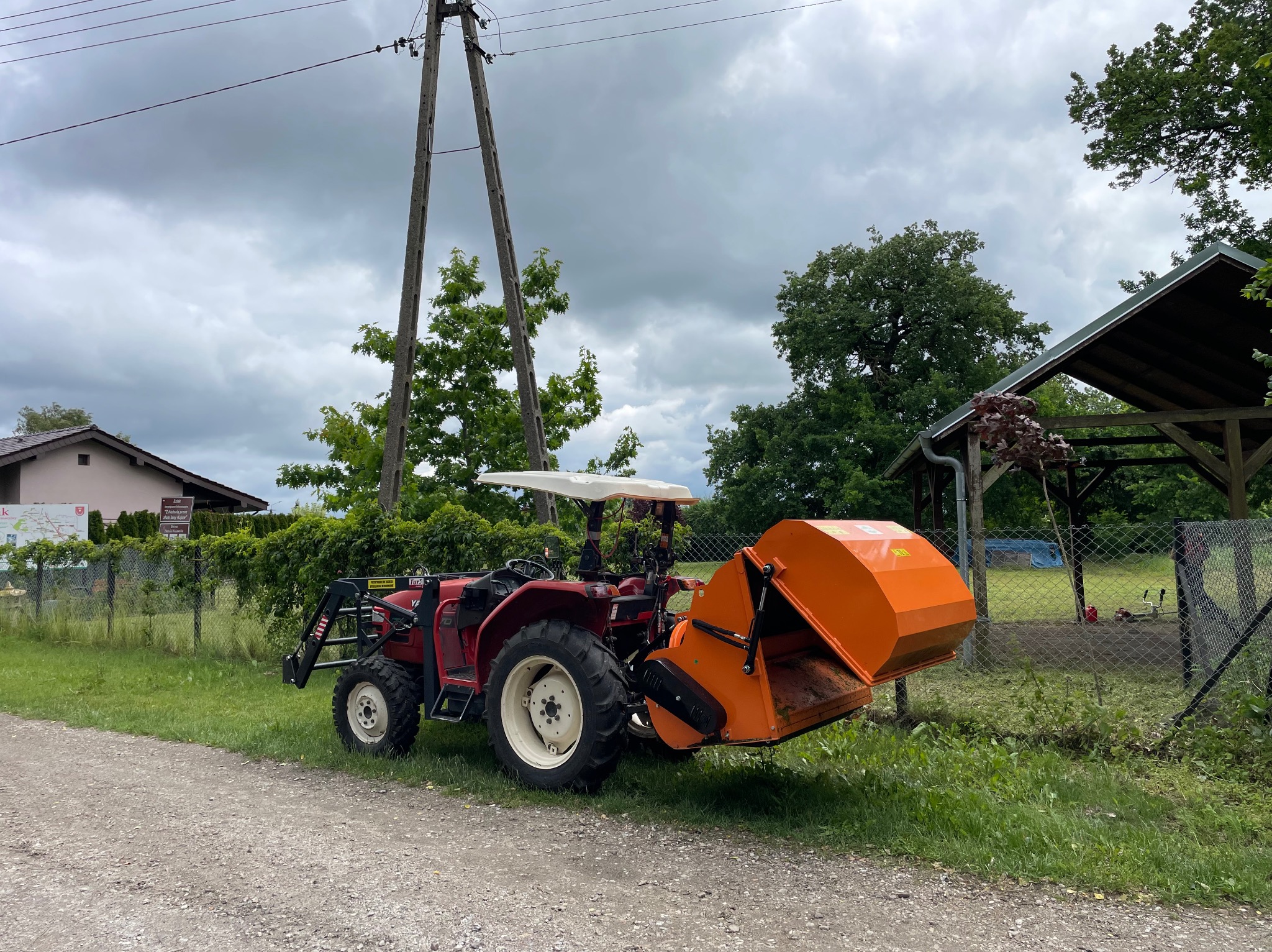 Czerwony traktor z pomarańczowym osprzętem na tle zieleni i pochmurnego nieba, gotowy do pracy w ogrodzie lub na działce. Widoczny słup energetyczny w tle.