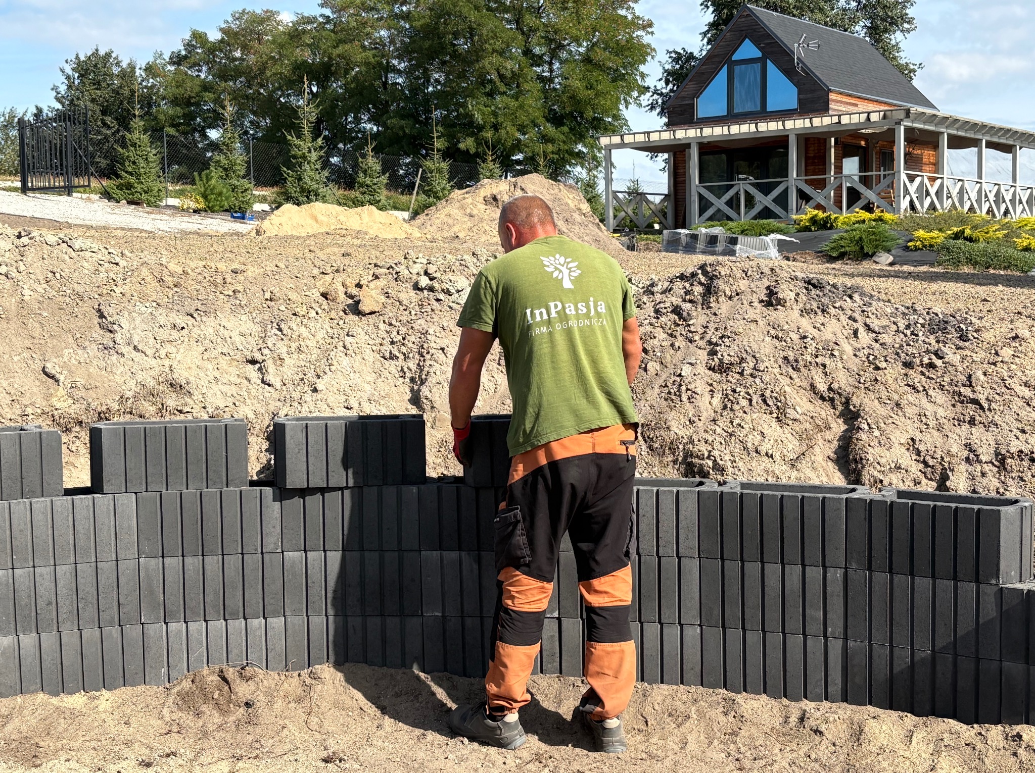 Układanie murku oporowego z betonowych bloczków przez pracownika w zielonej koszulce z logo firmy ogrodniczej, w tle dom i przygotowany teren pod ogród.