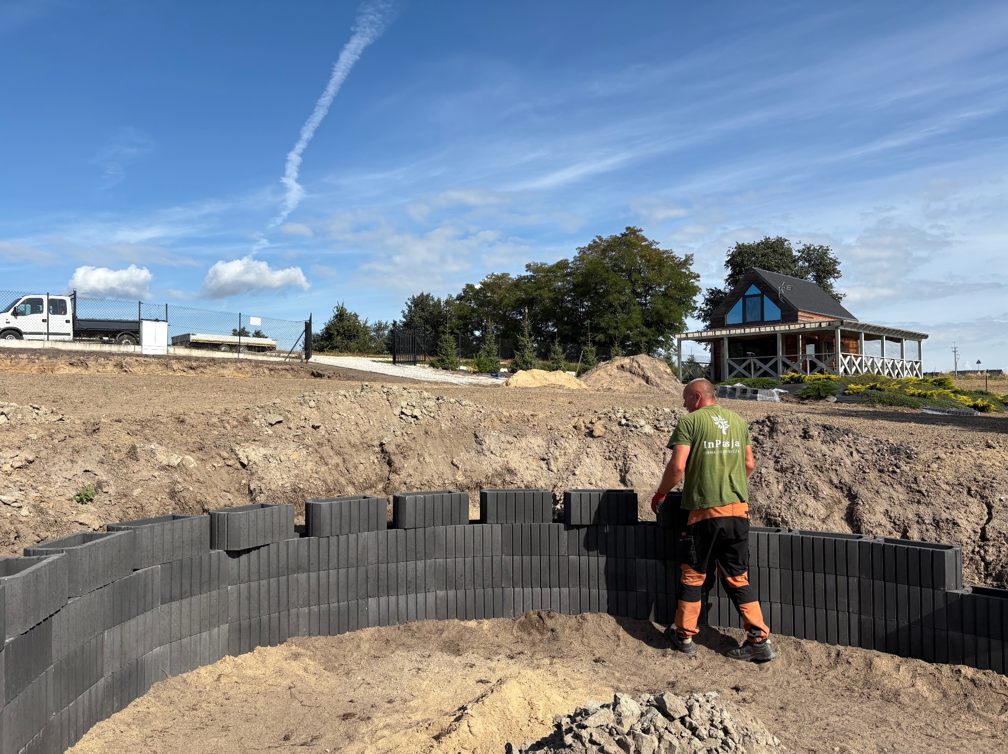 Budowa murku oporowego z szarych bloczków betonowych na piaszczystym podłożu. W tle dom z drewnianą elewacją i zielenią. Pracownik w odzieży roboczej. Błękitne niebo.