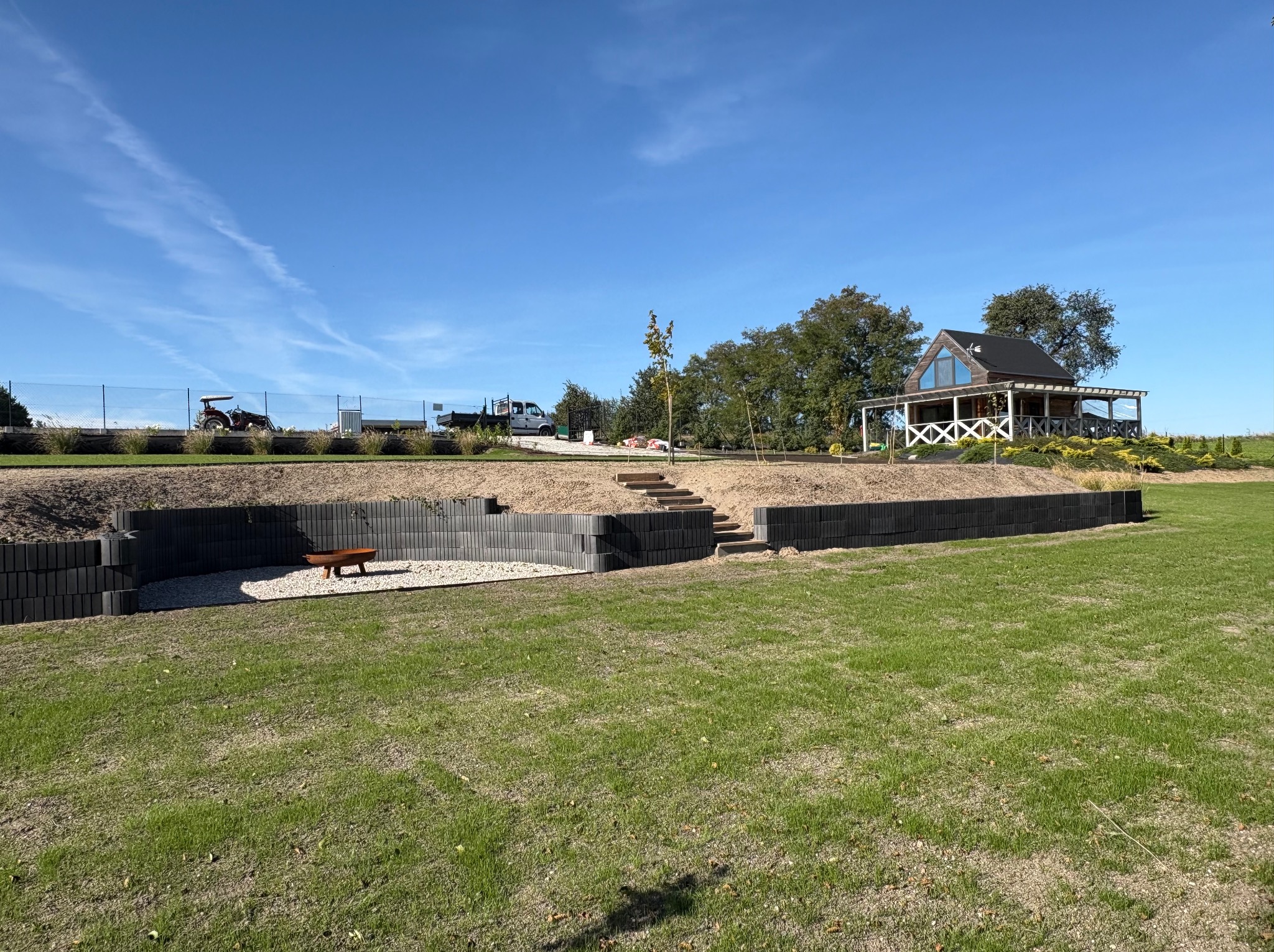 Ogród po realizacji w Kruszwicy: Murki oporowe z kostki, schody terenowe, trawnik i mała architektura. Widok na dom z werandą i błękitne niebo.
