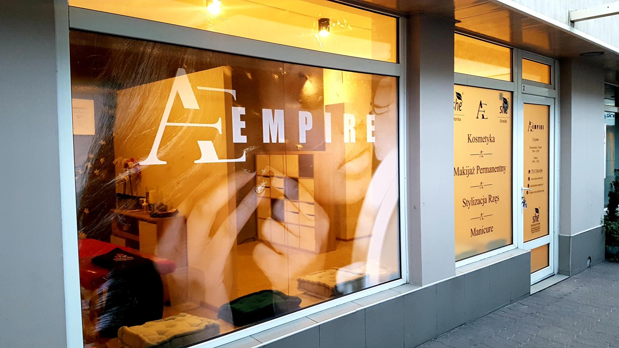 Witryna salonu kosmetycznego 'Empire' z naklejonym logo i ofertą usług: kosmetyka, makijaż permanentny, stylizacja rzęs, manicure. W tle widoczne wnętrze salonu z leżanką i półkami.