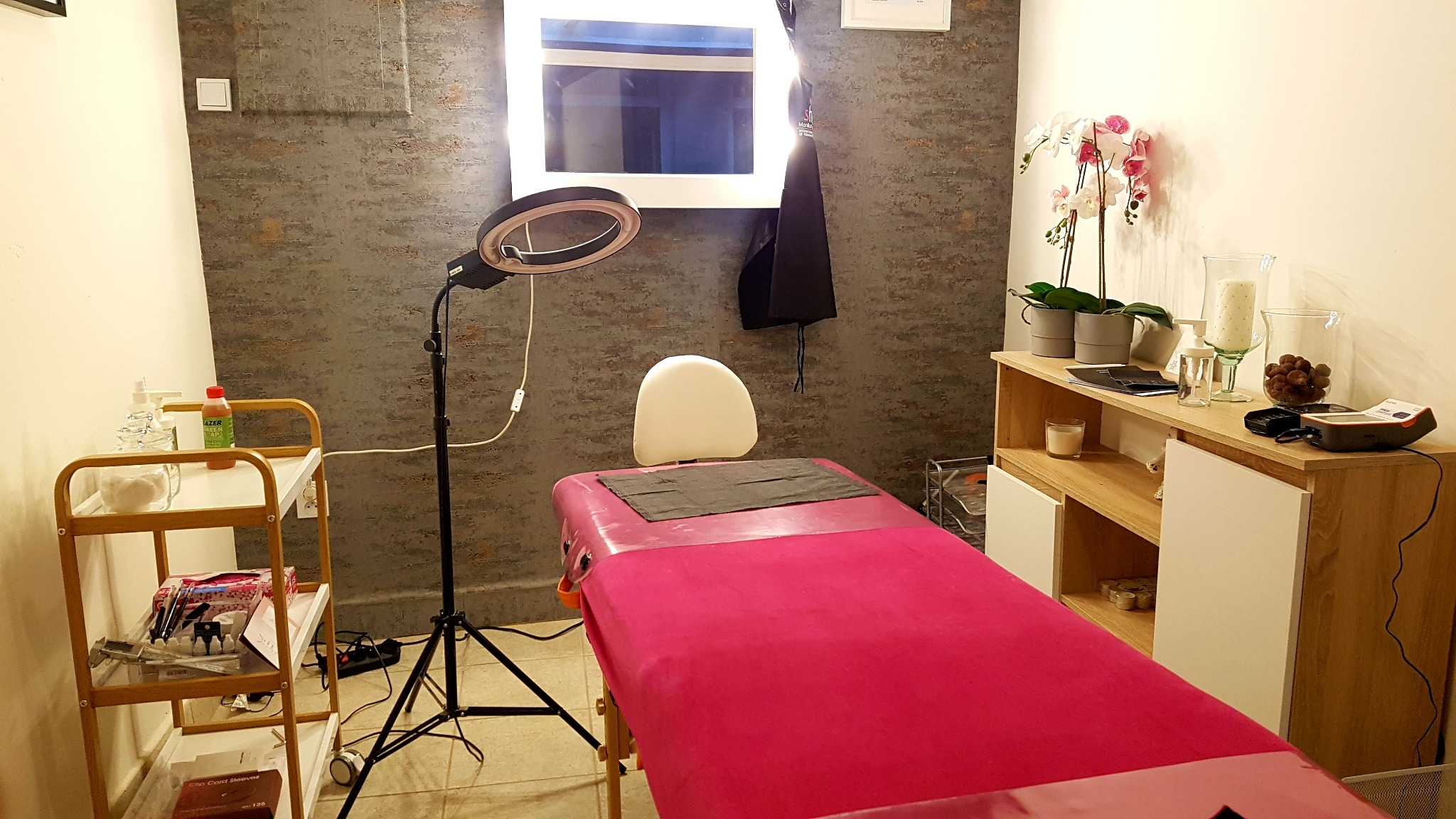 Wnętrze gabinetu z fotelem kosmetycznym przykrytym różowym materiałem, lampą pierścieniową, półką z kosmetykami i dekoracjami, ściana z efektem betonu.