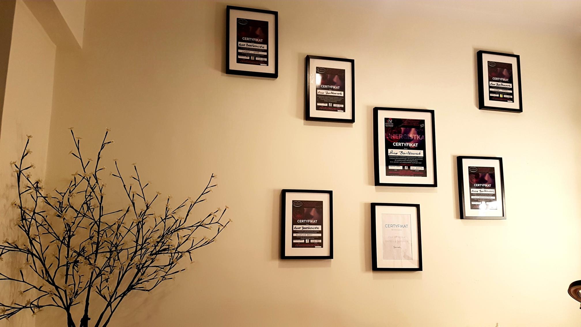 Ściana w gabinecie z powieszonymi certyfikatami w czarnych ramkach, widoczna dekoracyjna gałązka z białymi kwiatkami w wazonie.