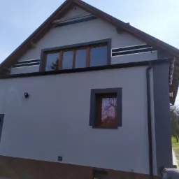 Elewacja domu z dwoma oknami z brązowymi ramami, wykończona w odcieniach szarości z dekoracyjnymi czarnymi pasami pod dachem, z widocznym fragmentem pracującego mężczyzny i wiadrem farby u dołu kadru.