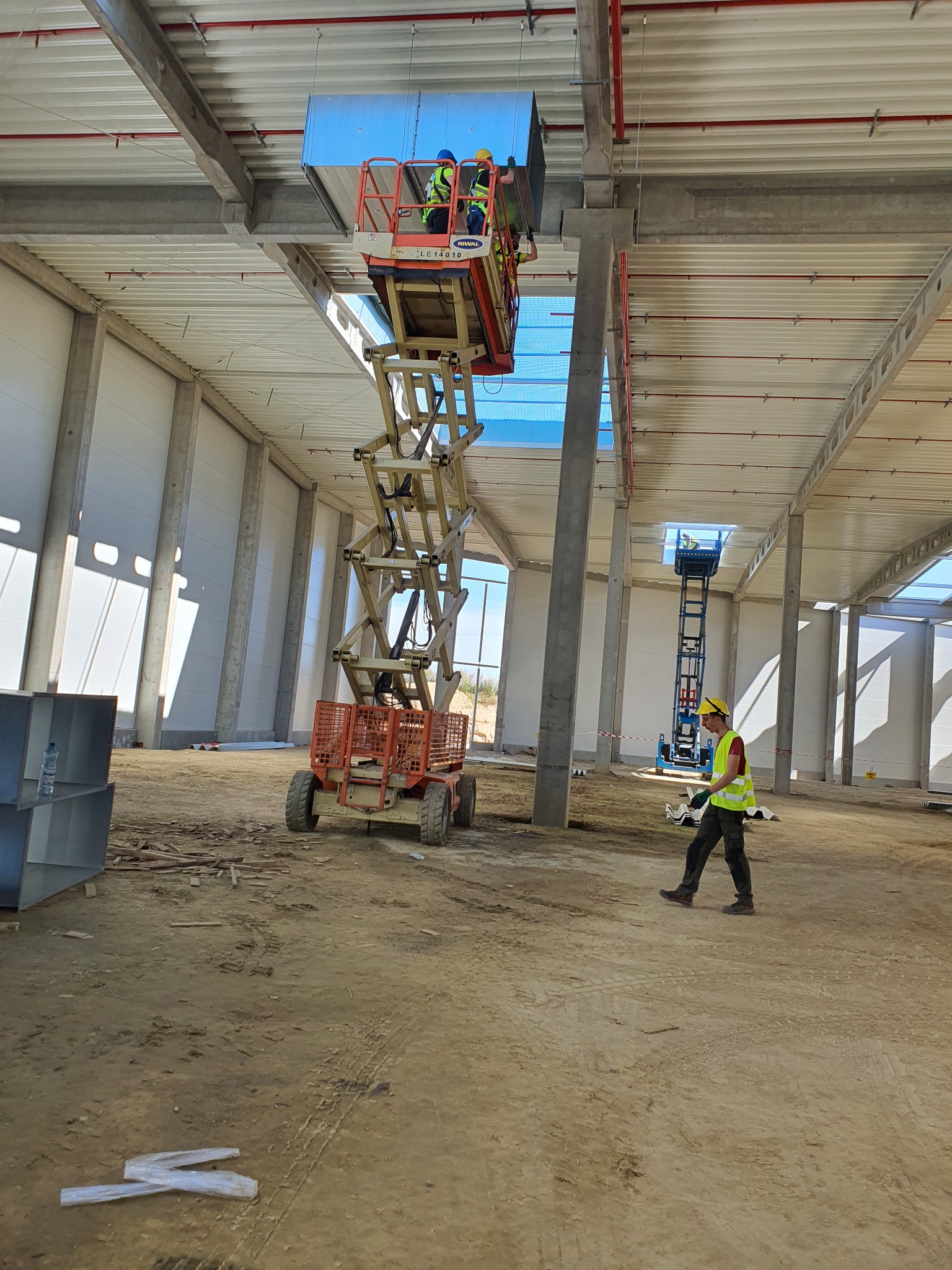 Dwóch monterów w podnośniku koszowym instaluje kanał wentylacyjny pod sufitem hali przemysłowej, pracownik w kamizelce odblaskowej nadzoruje prace z dołu.