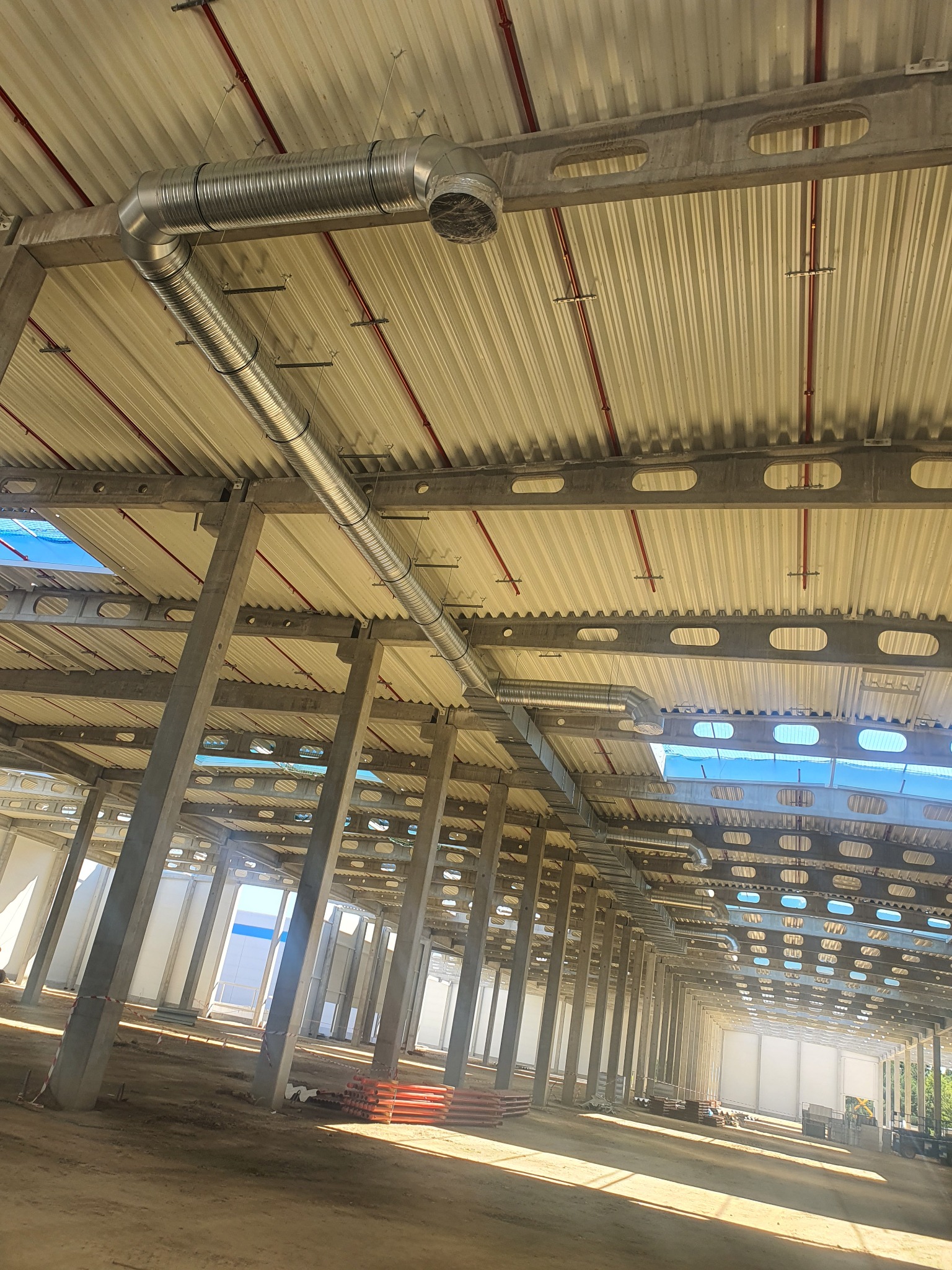 Wnętrze hali w trakcie budowy z widoczną stalową konstrukcją stropu i kolumnami, z zamontowanym systemem kanałów wentylacyjnych, widoczne świetliki w dachu, ziemia na podłodze.