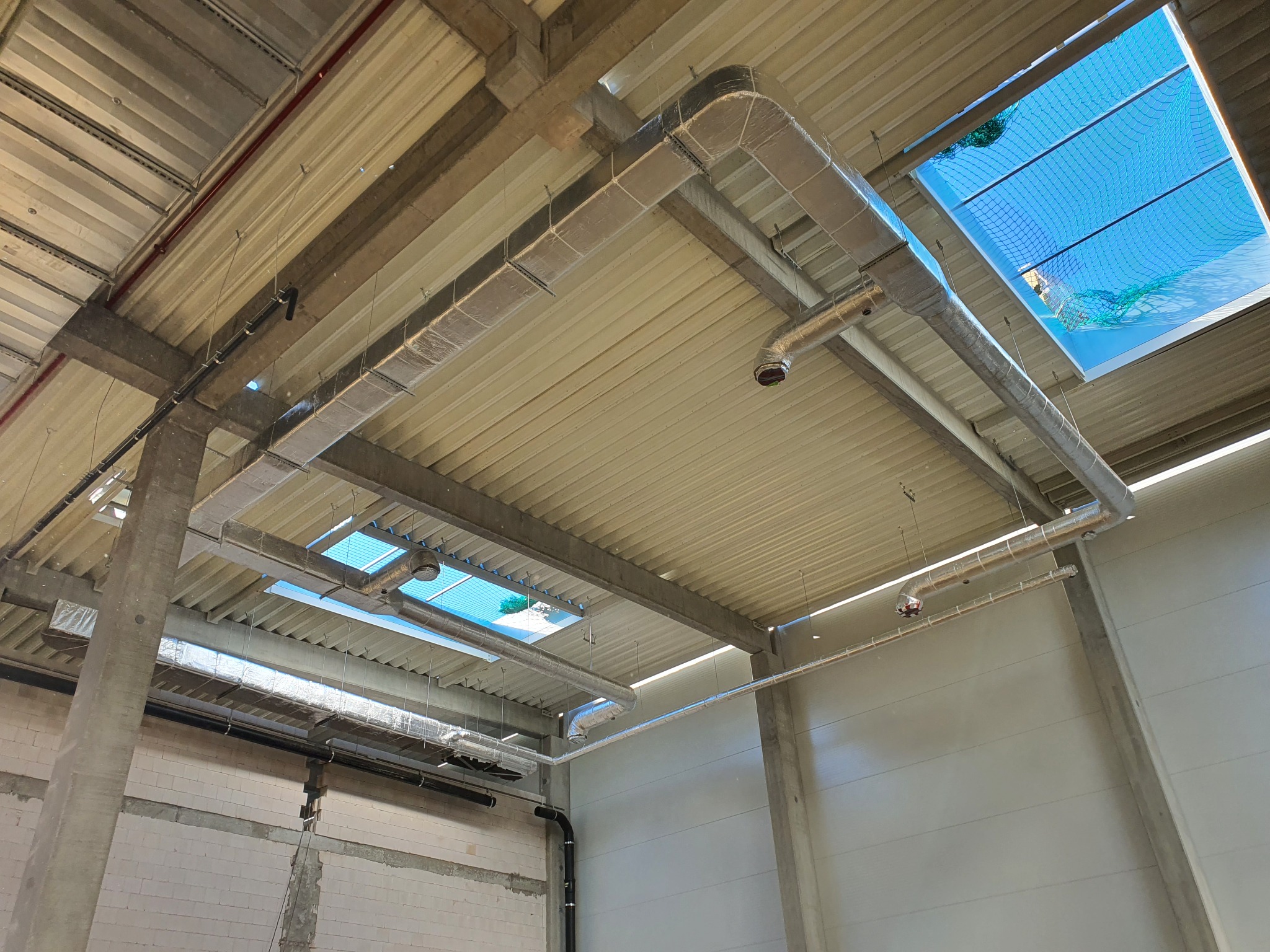 Szare kanały wentylacyjne pod sufitem hali przemysłowej, widoczne elementy mocujące, świetliki w suficie z niebieską siatką zabezpieczającą.