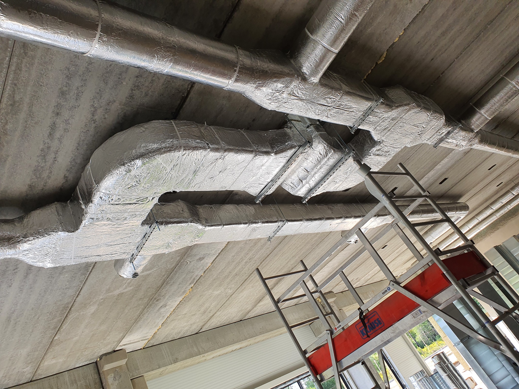 Izolowane, srebrną folią przewody wentylacyjne pod sufitem z betonu, widoczne z dołu, z fragmentem rusztowania Krause po prawej stronie.
