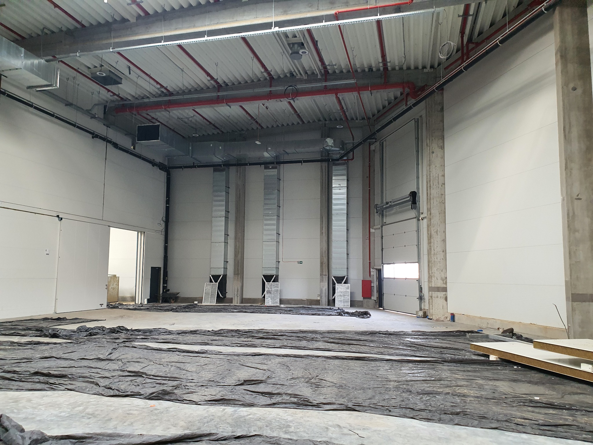 Wnętrze hali przemysłowej w trakcie montażu systemu wentylacji, widoczne pionowe kanały wentylacyjne przy ścianie, zabezpieczona folią podłoga i elementy instalacji pod sufitem.