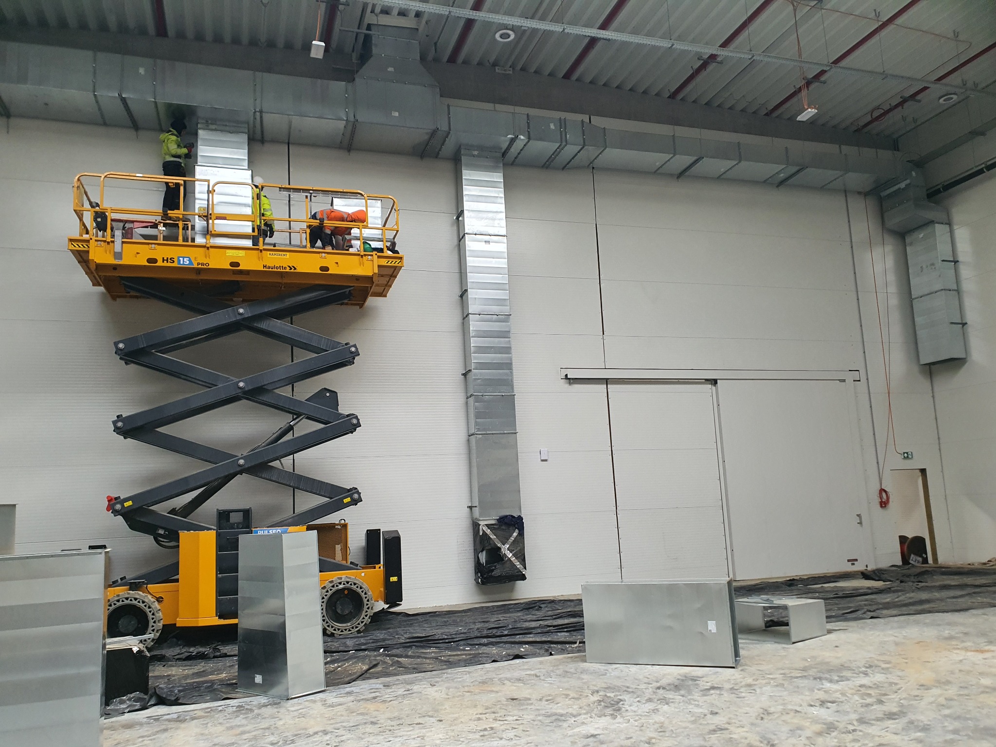 Montaż pionowego kanału wentylacyjnego z blachy ocynkowanej w hali przemysłowej, pracownicy na podnośniku koszowym, widoczne elementy instalacji pod sufitem.