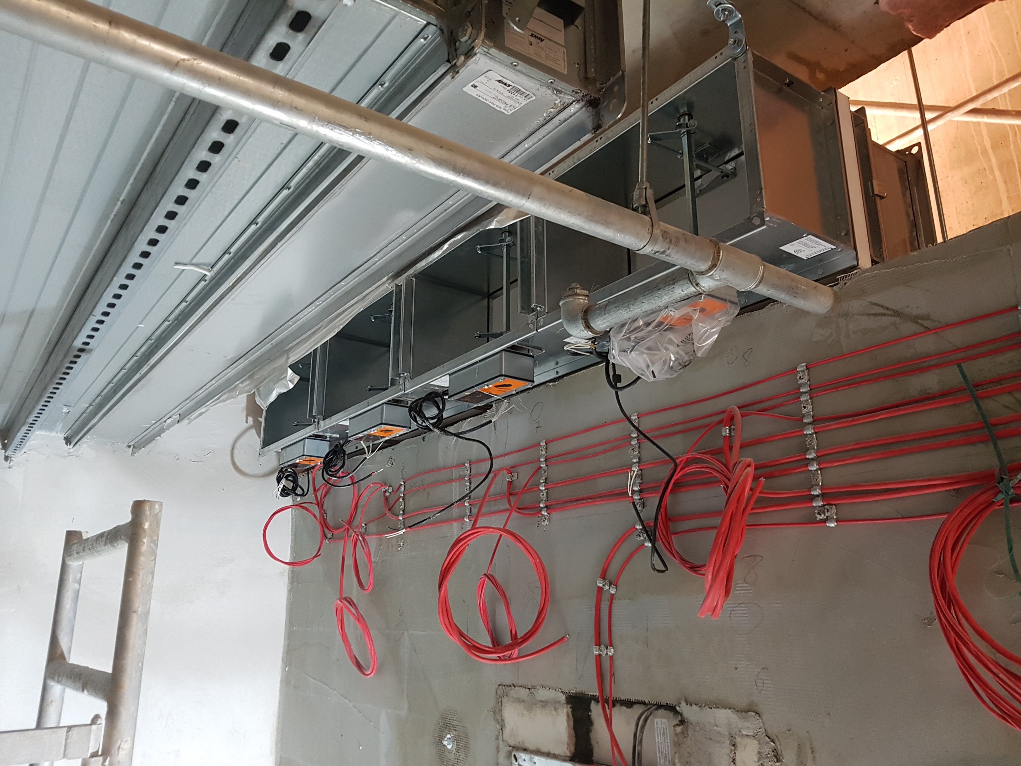 Metalowe kanały wentylacyjne pod sufitem w trakcie instalacji, widoczne czerwone przewody elektryczne i drabina aluminiowa.