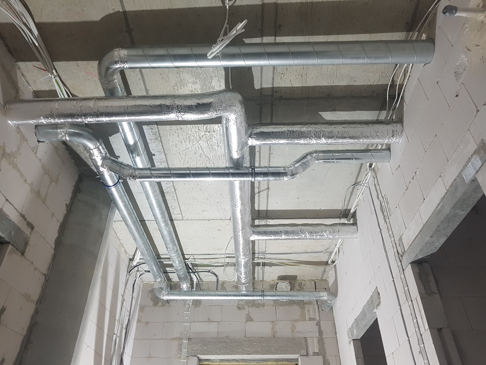 Montaż instalacji wentylacyjnej w surowym stanie budynku z widocznymi przewodami wentylacyjnymi, niektóre z izolacją, oraz przewodami elektrycznymi pod sufitem.