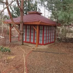 Altana w stylu japońskim z czerwoną dachówką i ścianami z przesuwanych paneli, otoczona drzewami i ściółką z kory.