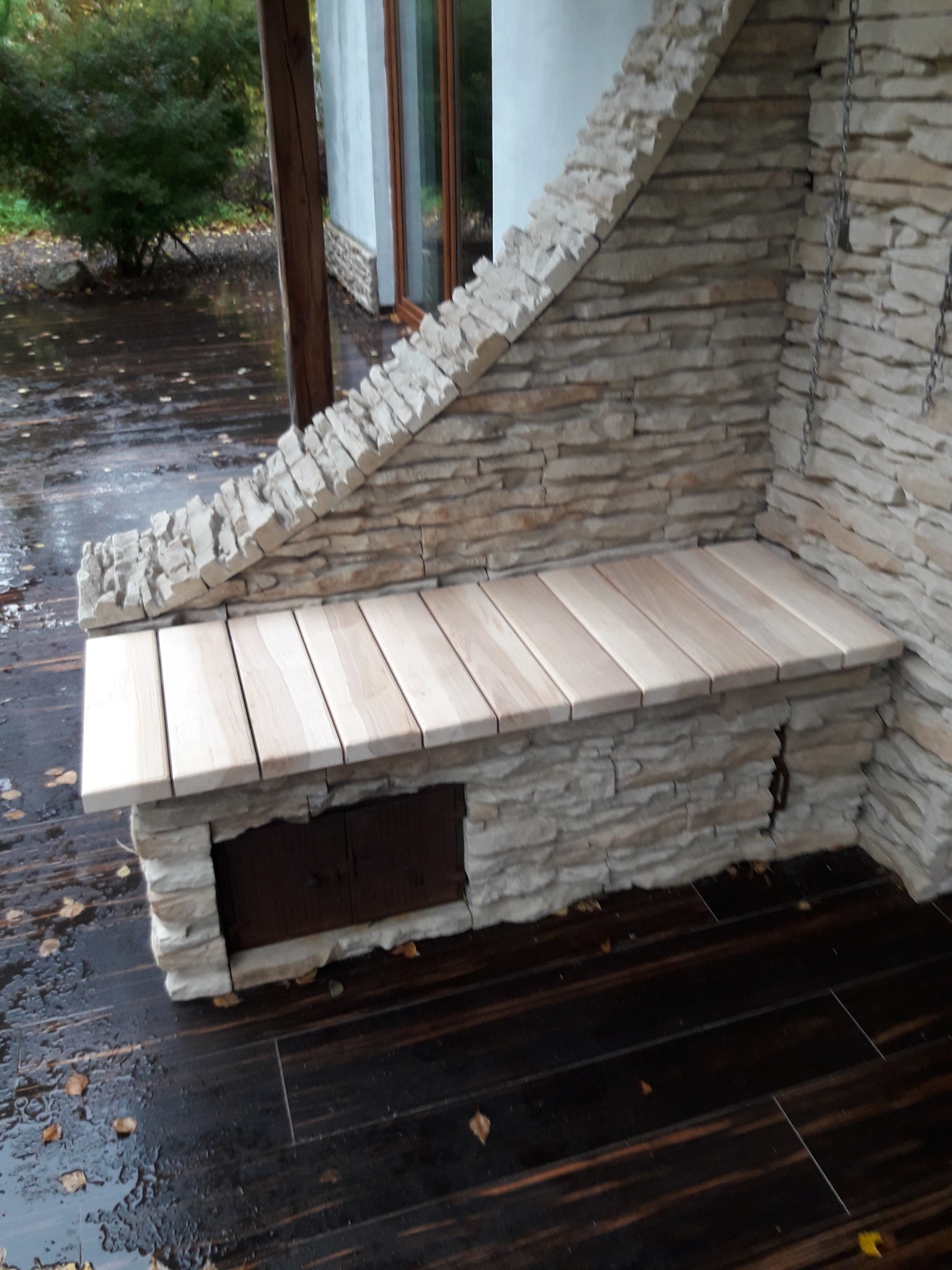 Kamienna ławka z drewnianym siedziskiem na zewnątrz, z małym schowkiem pod spodem, otoczona mokrym, ciemnym drewnianym tarasem i jesiennymi liśćmi.