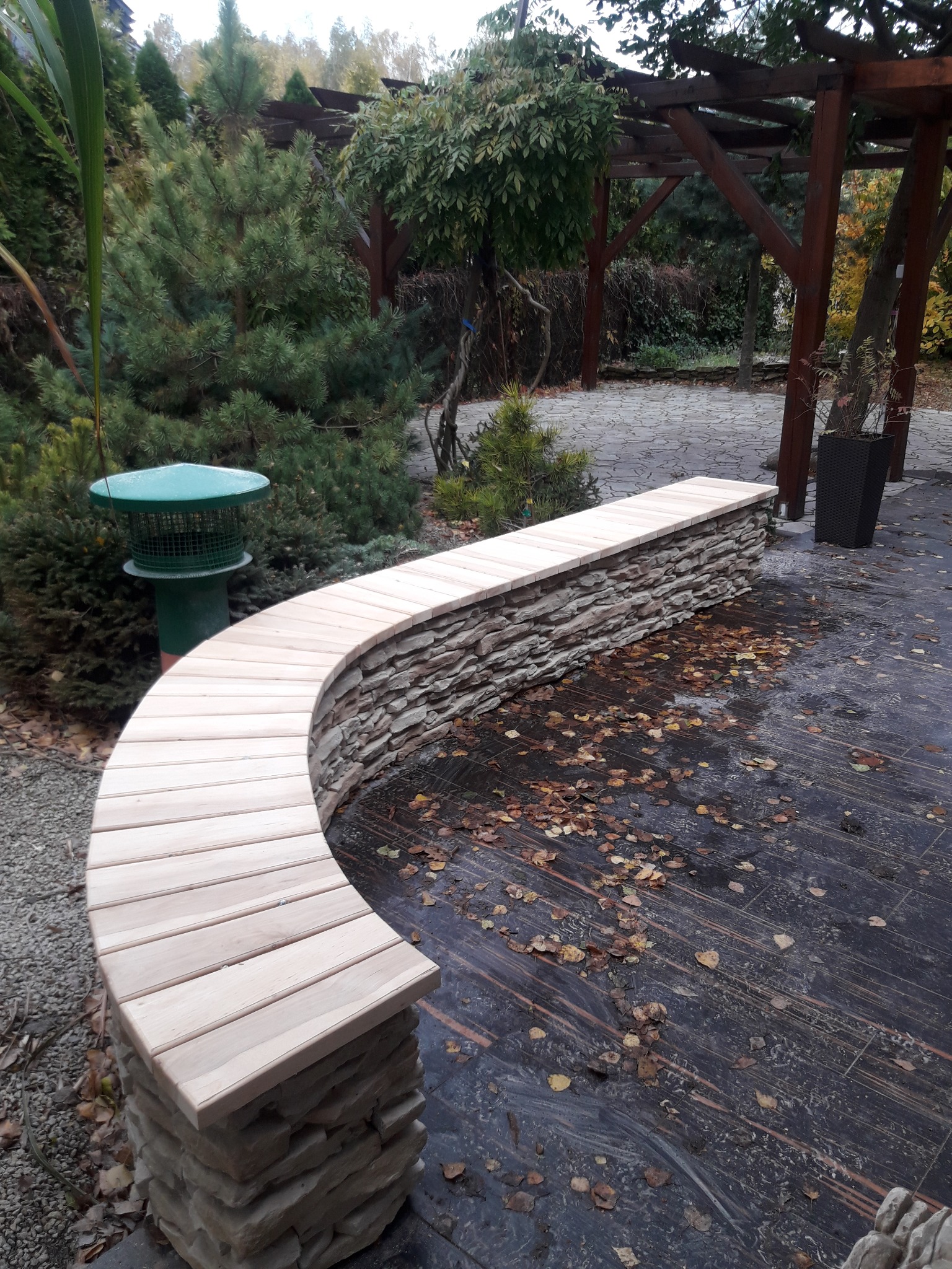 Zakrzywiona ławka z drewnianym siedziskiem i kamienną podstawą w ogrodzie, otoczona zielenią i opadłymi liśćmi na ciemnej podłodze.