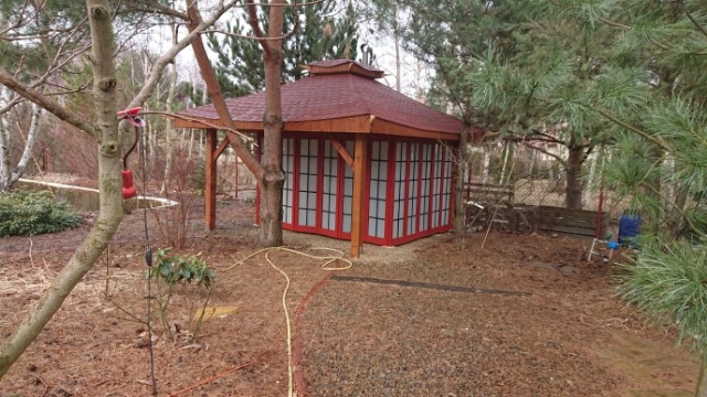 Altana w stylu japońskim z czerwoną dachówką i ścianami z przesuwanych paneli, otoczona drzewami i ściółką z kory.