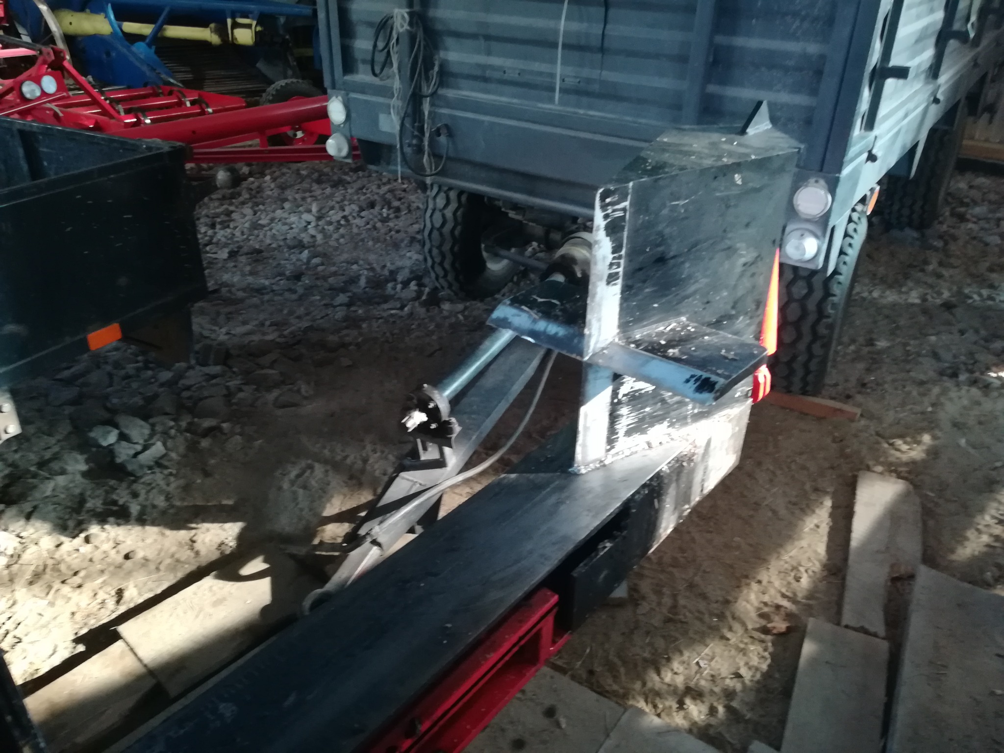 Fragment przyczepy z elementami metalowymi, widoczne spawane łączenia i fragmenty konstrukcji w kolorze czarnym i czerwonym, w tle koła przyczepy i gruz.