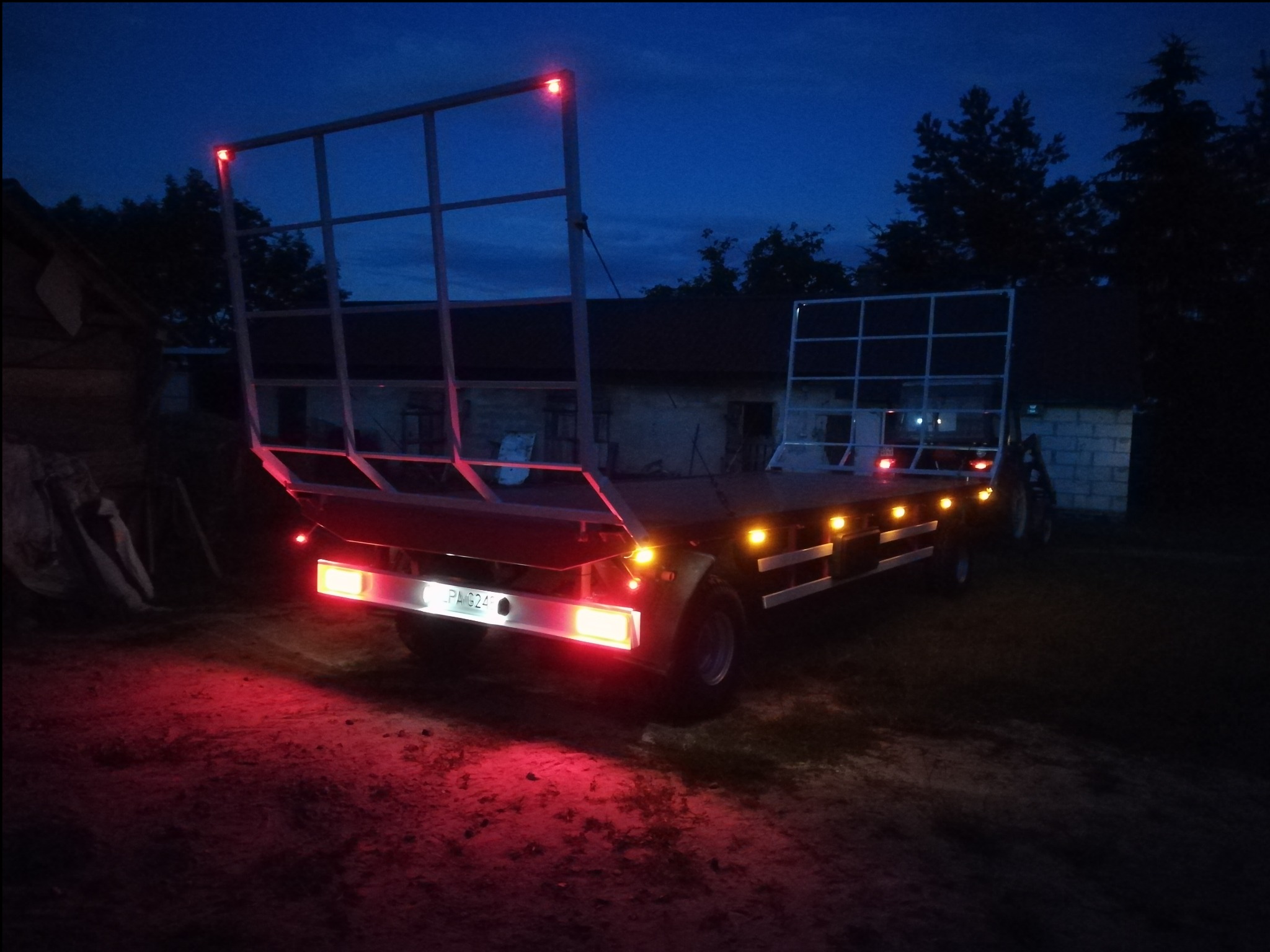 Przyczepa rolnicza z metalową konstrukcją zabezpieczającą ładunek, oświetlona czerwonymi lampami w nocy.