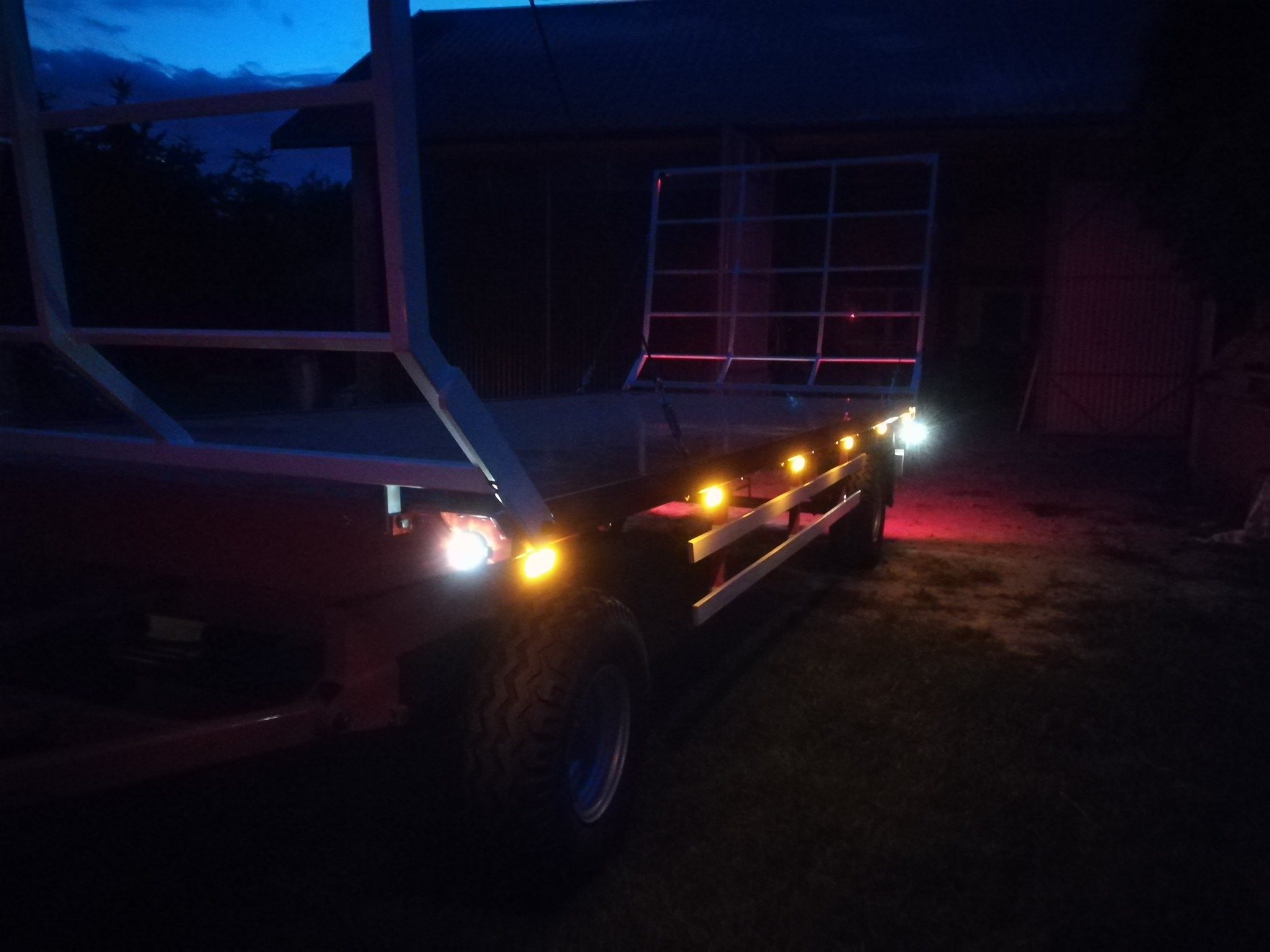 Przyczepa rolnicza z metalową konstrukcją skrzyni, oświetlona światłami pozycyjnymi w ciemności, widoczna opona z bieżnikiem.