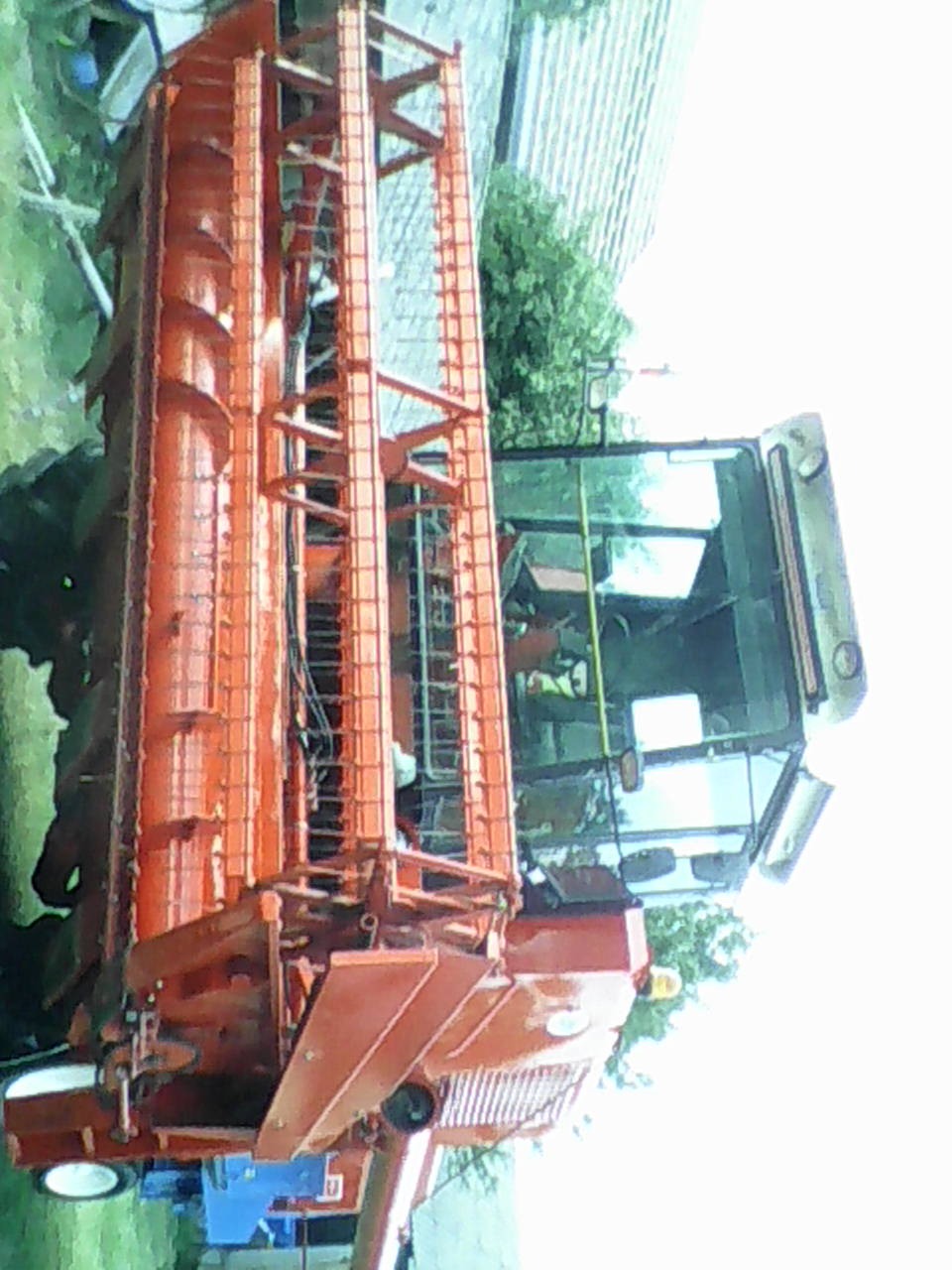 Pomarańczowa maszyna rolnicza, prawdopodobnie kombajn, z widocznymi elementami żniwnymi i kabiną operatora, sfotografowana w słoneczny dzień na tle zieleni.