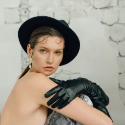 Portret modelki z Wrocławia w czarnym kapeluszu i długich skórzanych rękawiczkach, pozującej na tle ściany z białej cegły.