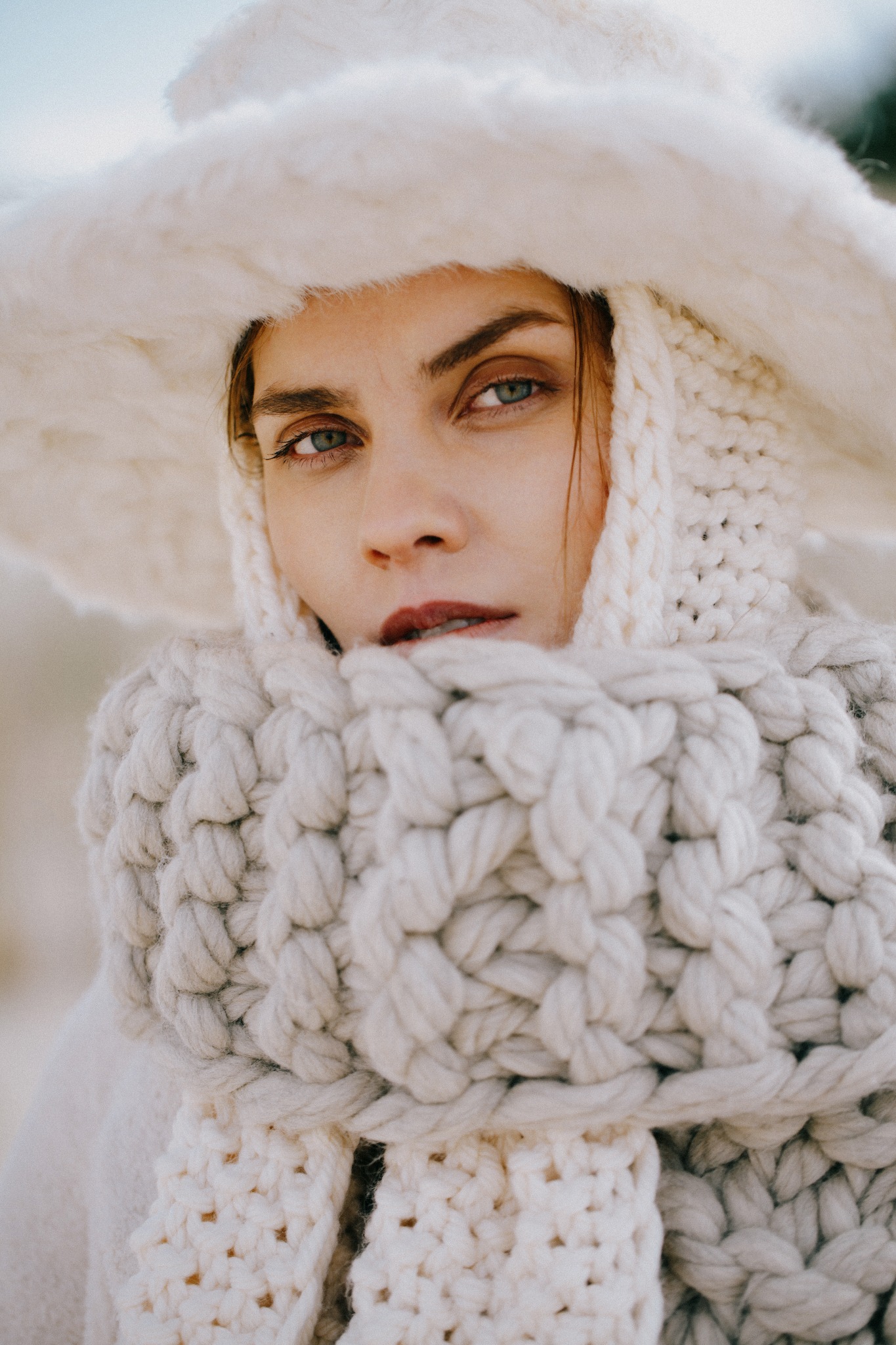 Portret kobiety w obszernym, kremowym szalu z grubych splotów i futrzanym kapeluszu, makijaż z ciemną szminką, spojrzenie skierowane wprost.