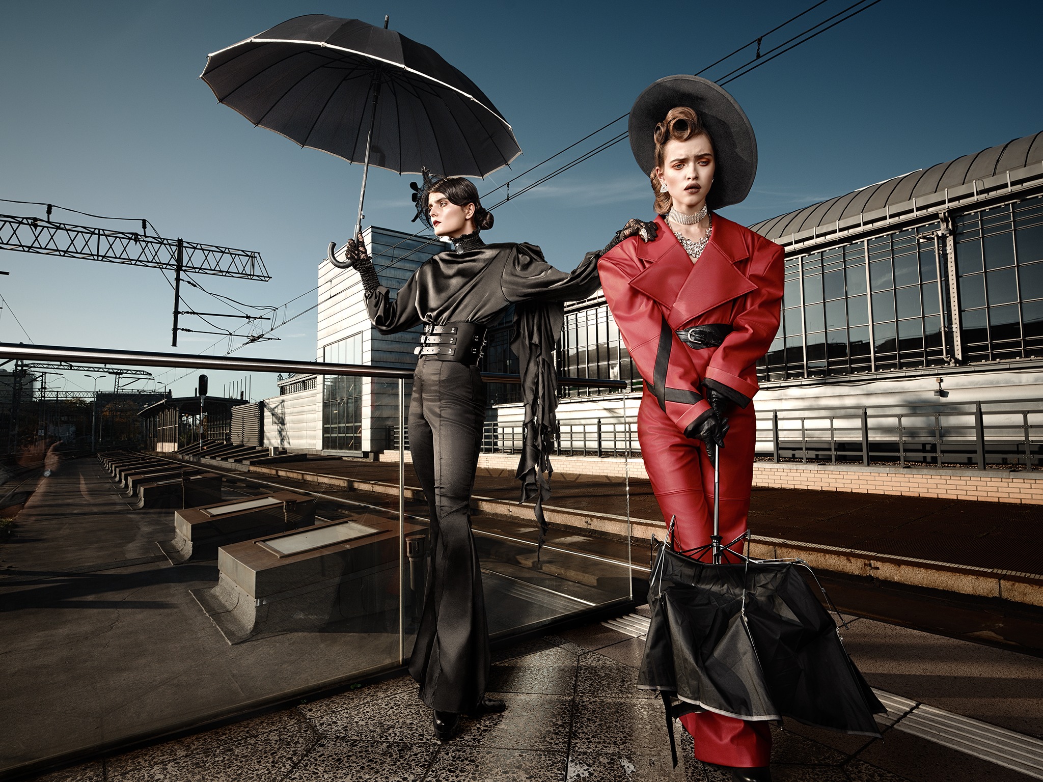 Dwie modelki w stylizowanych strojach na tle dworca: jedna w czarnym, satynowym komplecie z parasolem, druga w czerwonym, skórzanym płaszczu i spodniach, z dużym kapeluszem i parasolem.