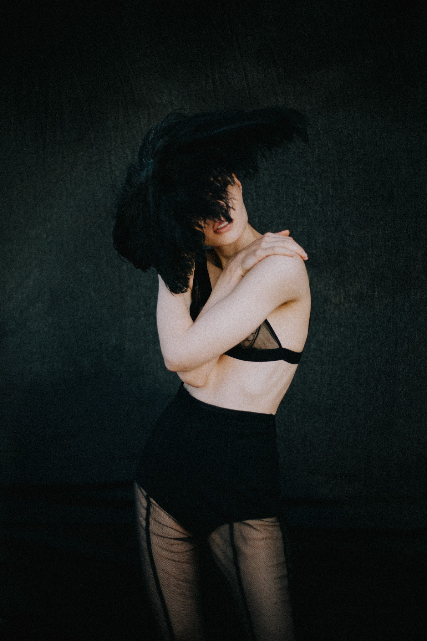 Kobieta w czarnej bieliźnie i kapeluszu z piór pozuje na tle ciemnej ściany, krzyżując ramiona.
