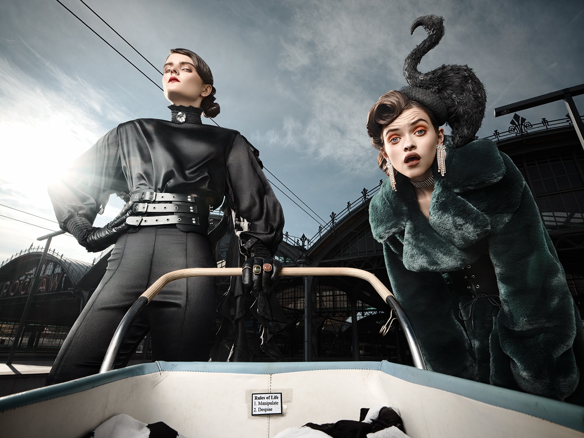 Dwie modelki w stylizowanych, awangardowych strojach na tle budynku dworca kolejowego; jedna w czarnym, obcisłym stroju z szerokim pasem, druga w zielonym futrze i z czarnym łabędziem na głowie...